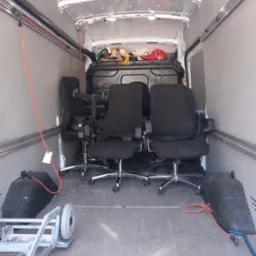 Wnętrze furgonetki z trzema czarnymi fotelami biurowymi zabezpieczonymi pasami, w tle widoczne przedmioty pod plandeką, w dolnym lewym rogu wózek transportowy.