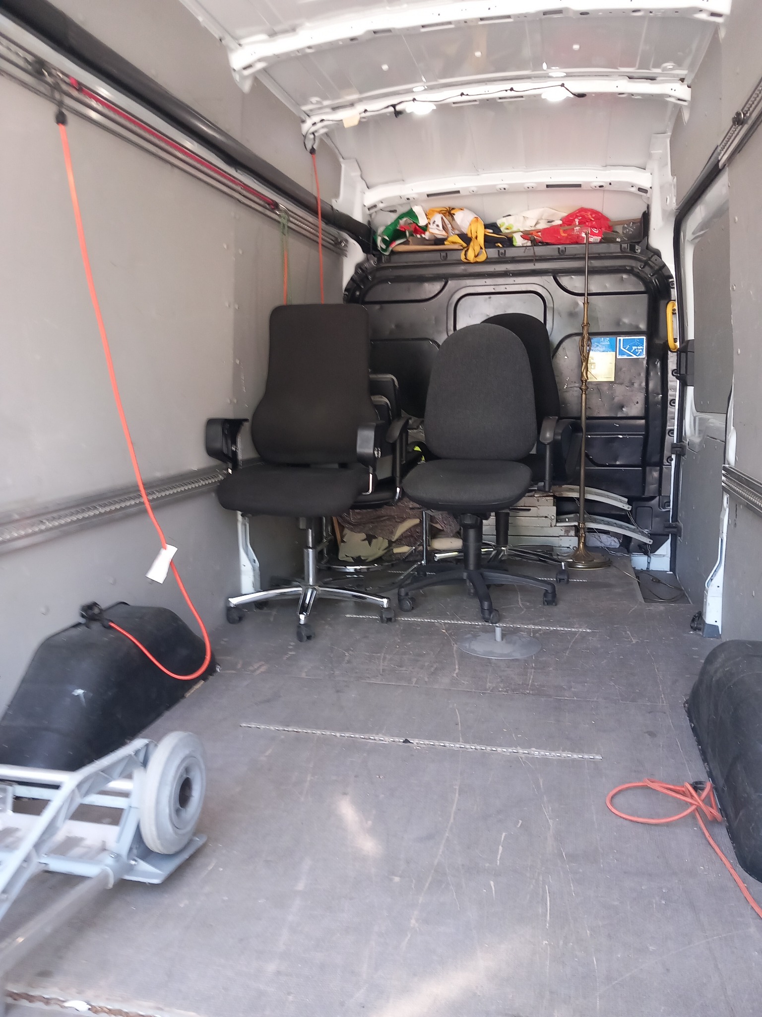Wnętrze furgonetki załadowane krzesłami biurowymi, lampą stojącą i wózkiem transportowym, zabezpieczone pasami w trakcie transportu.