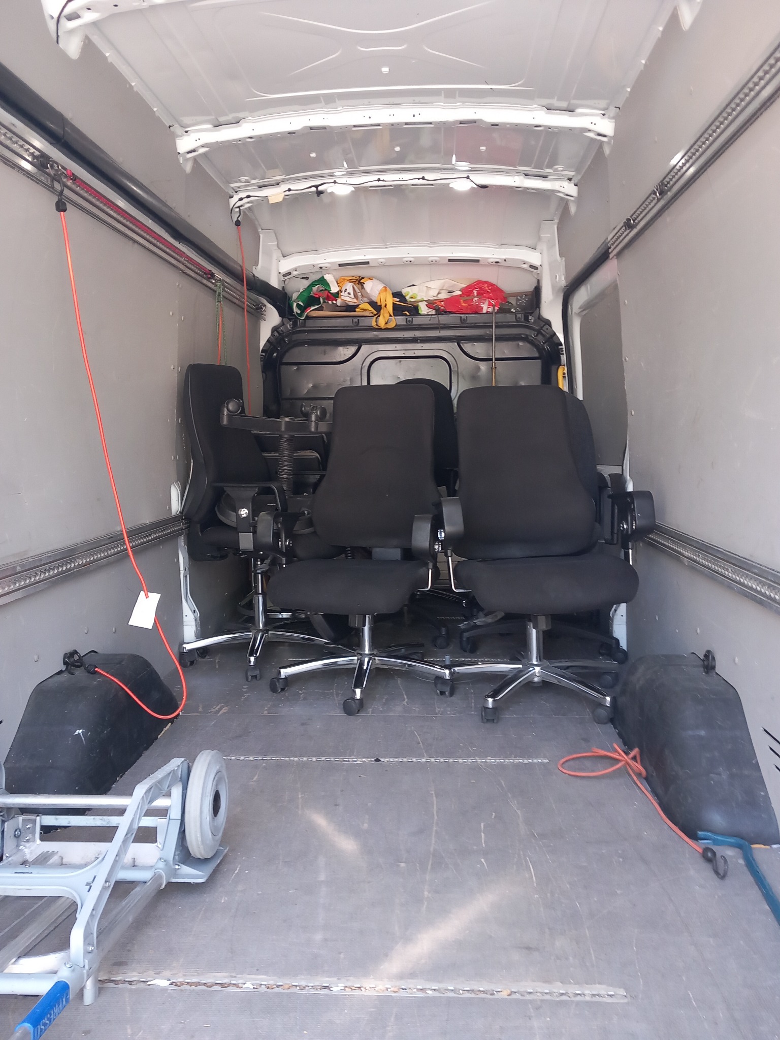 Wnętrze furgonetki z trzema czarnymi fotelami biurowymi zabezpieczonymi pasami, w tle widoczne przedmioty pod plandeką, w dolnym lewym rogu wózek transportowy.