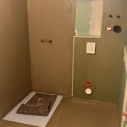 Wnętrze łazienki w trakcie prac wykończeniowych: ściany częściowo pokryte szarobrązową warstwą, widoczne instalacje hydrauliczne, podłoga zalana wylewką, zabezpieczona mata ochronną nad brodzikiem.