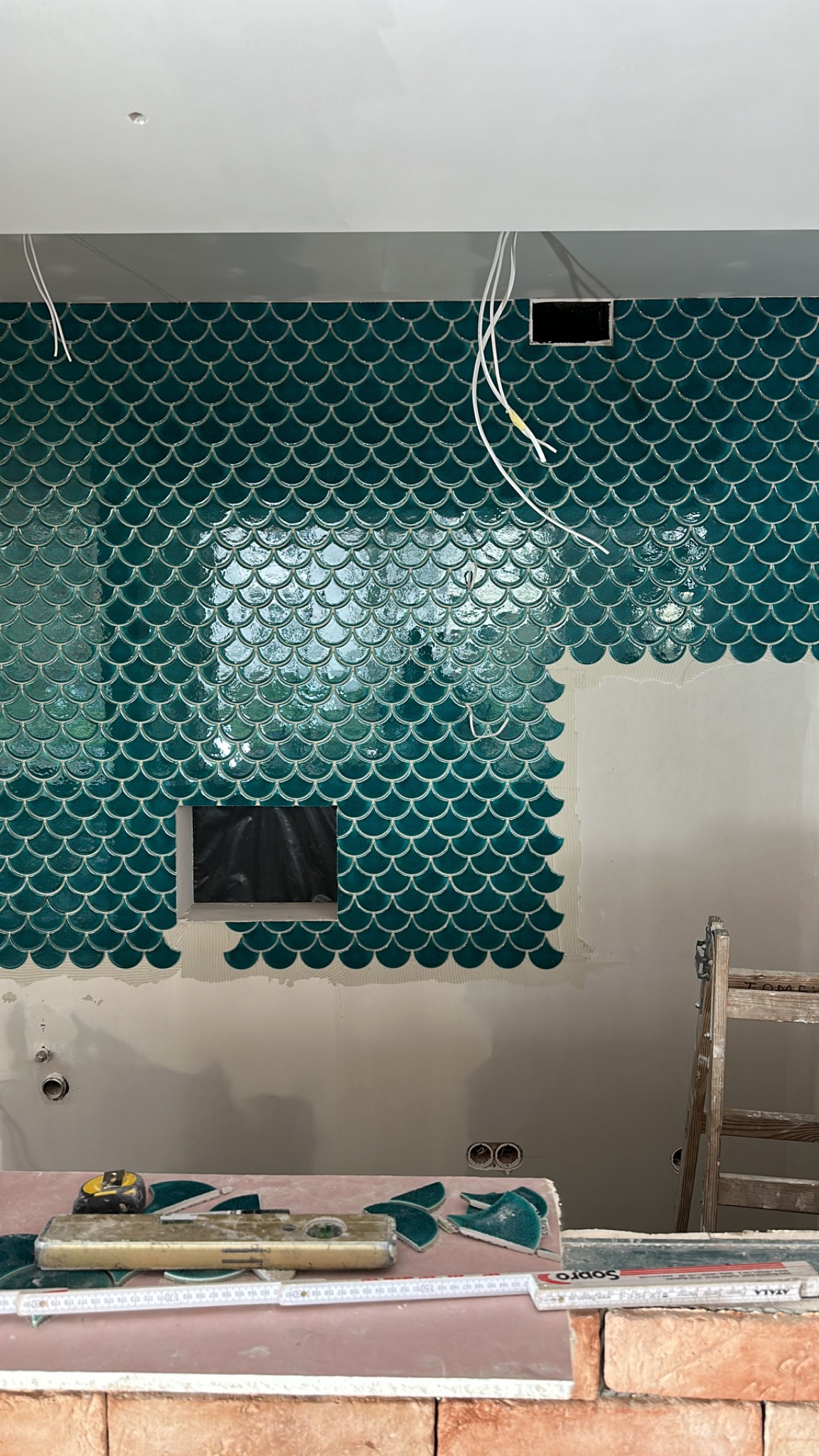 Układanie błyszczących, turkusowych płytek w kształcie rybiej łuski na ścianie w kuchni. Widoczne narzędzia, poziomica i fragmenty płytek na blacie z cegieł.