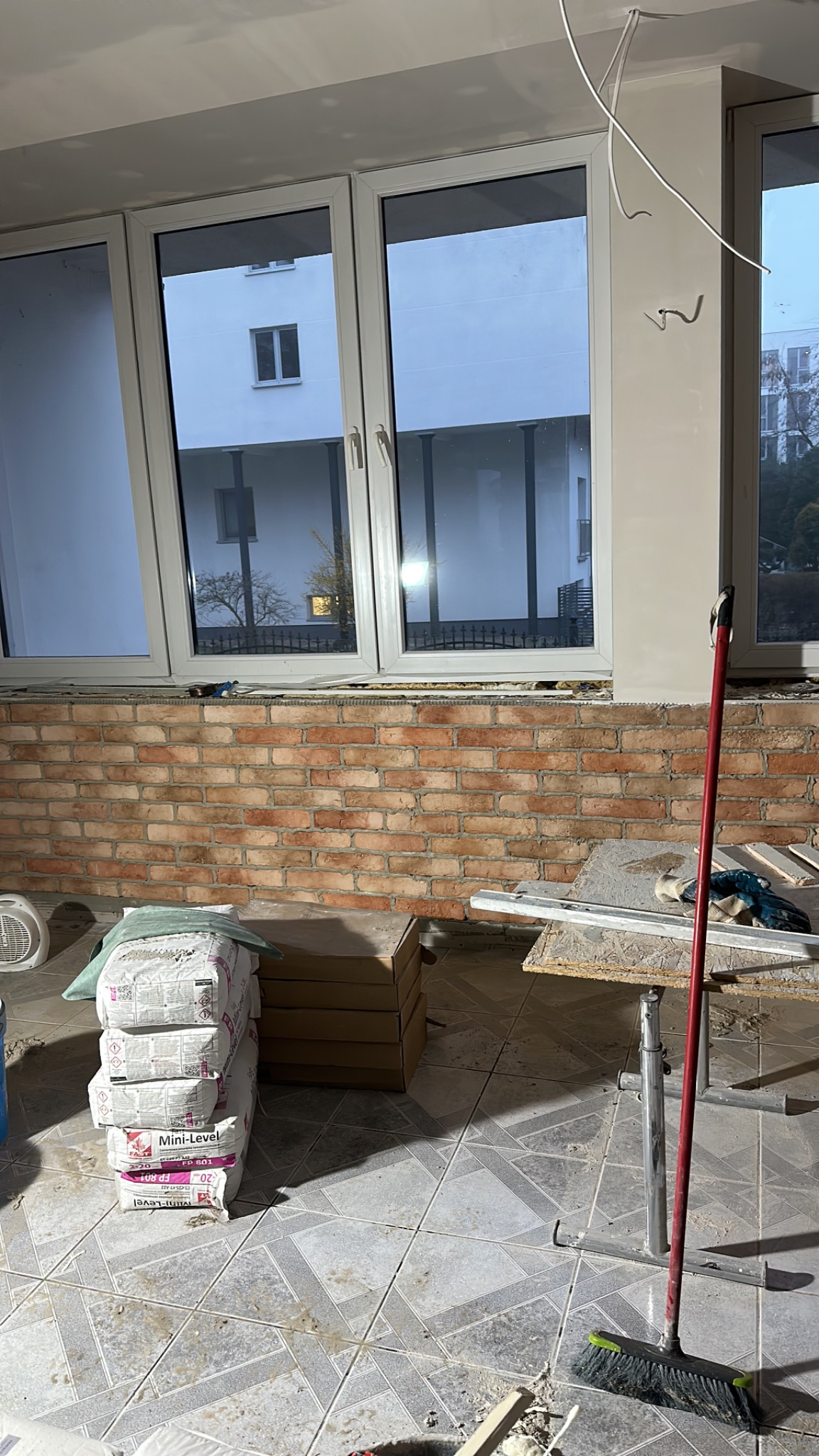 Wnętrze w trakcie prac murarskich: ściana z czerwonej cegły pod oknem, obok worki z zaprawą, drabina i narzędzia, widoczne kable wystające z sufitu.