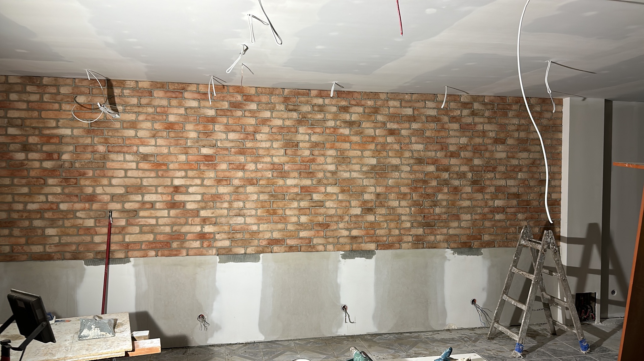 Ściana z cegły klinkierowej w trakcie montażu, dolna część ściany przygotowana pod tynk, widoczne wystające kable elektryczne, drabina i narzędzia.