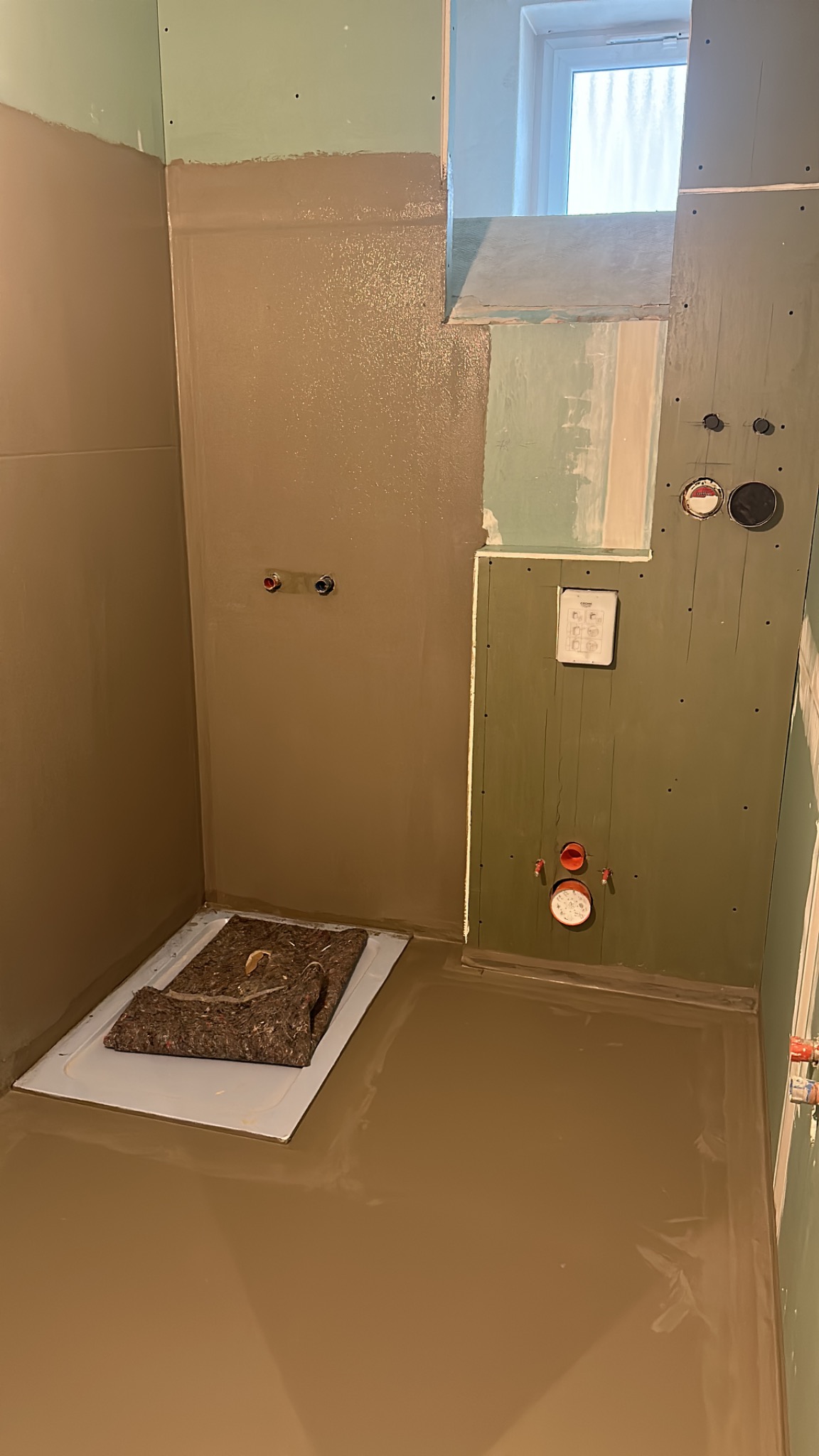 Wnętrze łazienki w trakcie prac wykończeniowych: ściany częściowo pokryte szarobrązową warstwą, widoczne instalacje hydrauliczne, podłoga zalana wylewką, zabezpieczona mata ochronną nad brodzikiem.