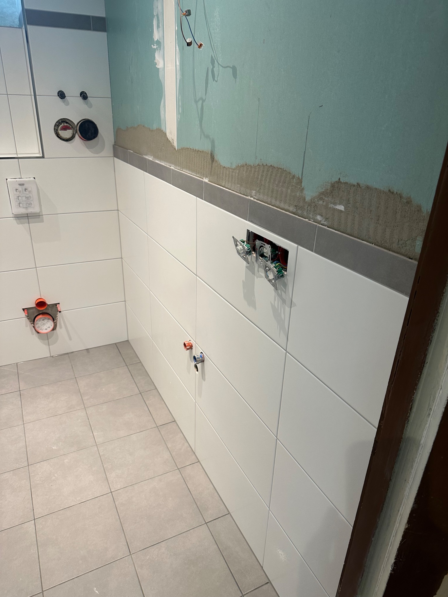 Wnętrze łazienki w trakcie remontu: białe płytki na ścianach ułożone do połowy wysokości, powyżej surowa ściana przygotowana pod tynk, widoczne instalacje elektryczne i hydrauliczne.