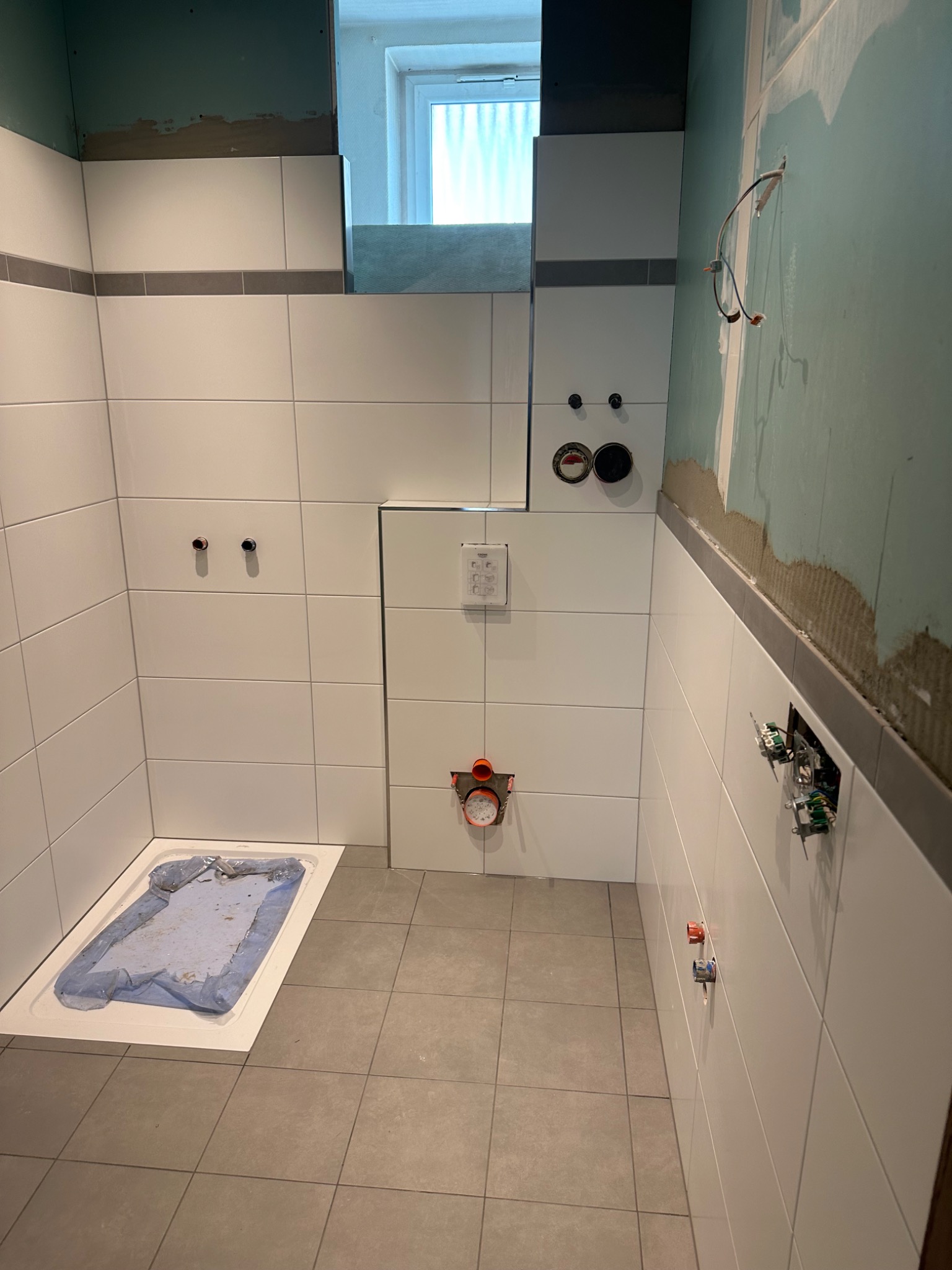 Wnętrze łazienki w trakcie prac wykończeniowych: białe płytki ścienne z szarym pasem, szare płytki podłogowe, zabezpieczony folią brodzik, odsłonięte przewody elektryczne i instalacje...