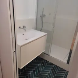 Nowoczesna łazienka z wiszącą szafką pod umywalką, kabiną prysznicową i podłogą wyłożoną ciemnymi płytkami w jodełkę.