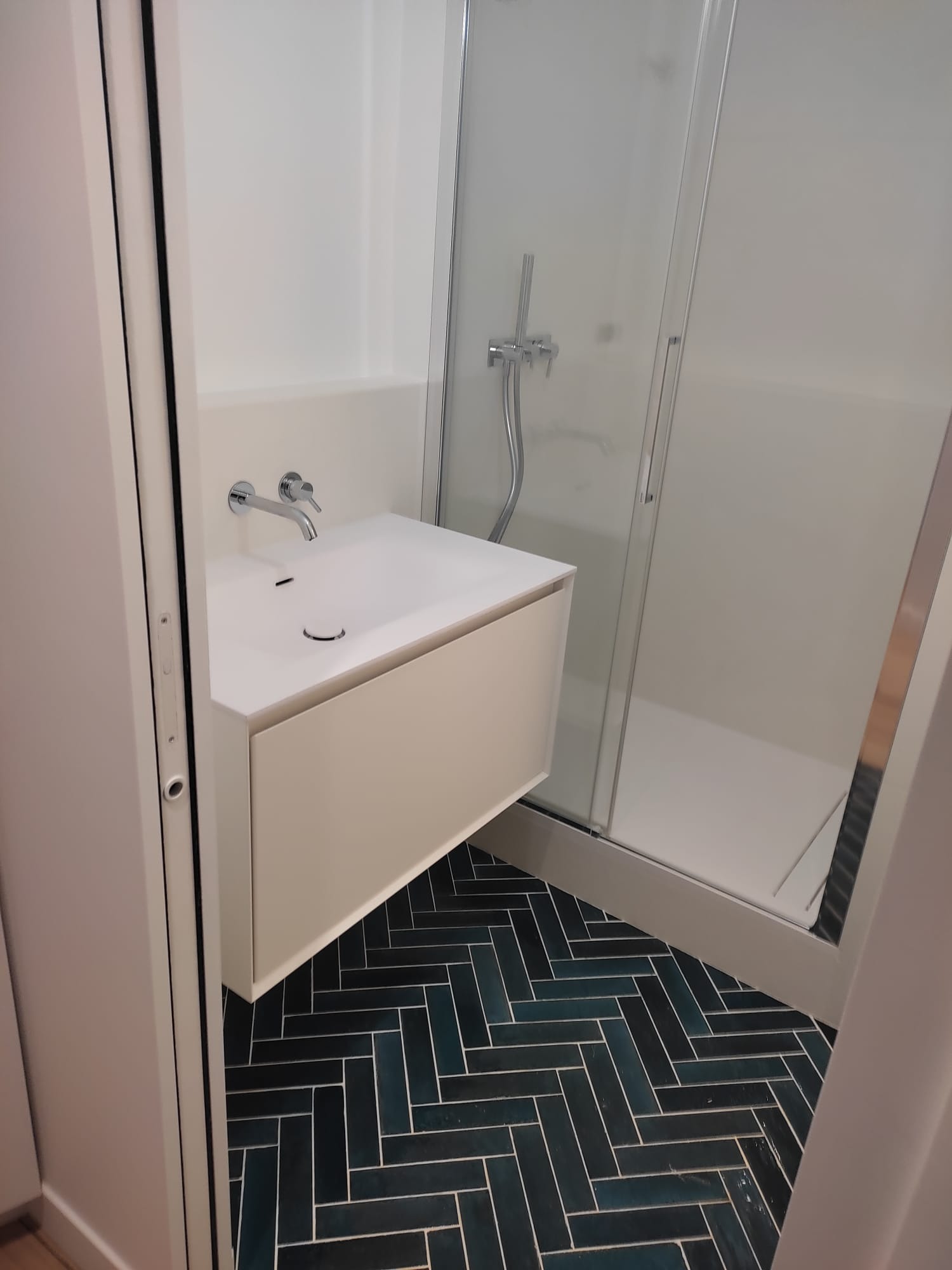 Nowoczesna łazienka z wiszącą szafką pod umywalką, kabiną prysznicową i podłogą wyłożoną ciemnymi płytkami w jodełkę.