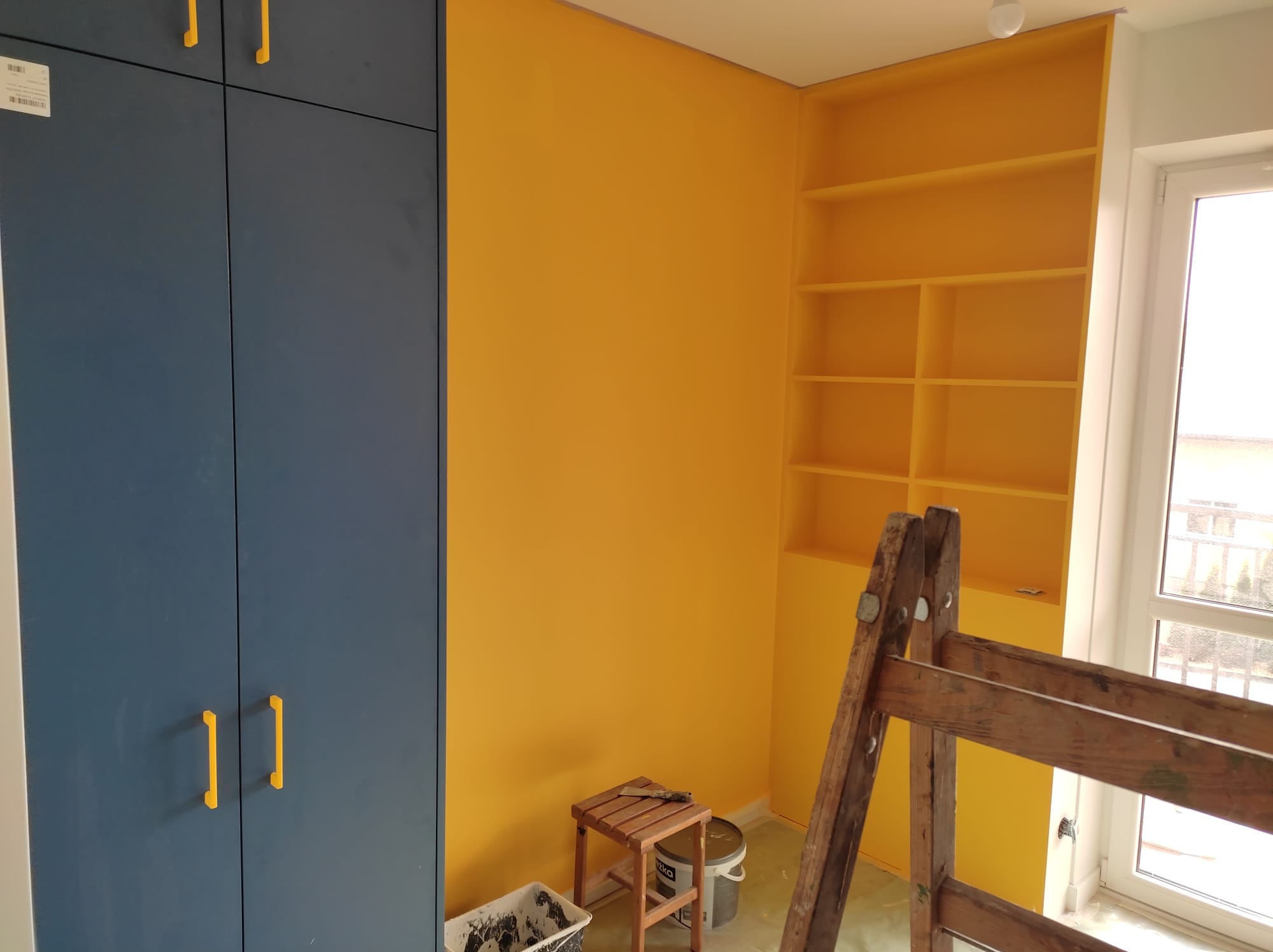 Pokój po odświeżeniu: ściana w intensywnym żółtym kolorze, drewniana drabina oparta o ścianę, niebieska szafa z żółtymi uchwytami po lewej stronie, widoczne wiaderka z farbą i stołek.