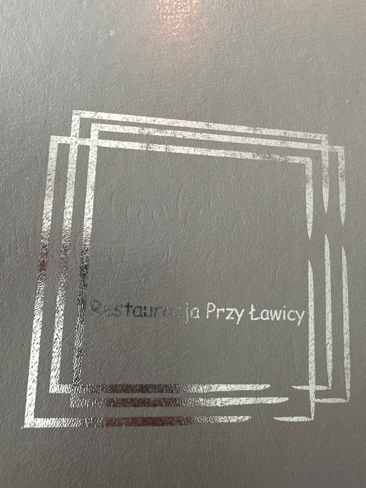 Szara okładka z tłoczonym srebrnym logo restauracji 'Restauracja Przy Ławicy' w geometrycznej ramce.