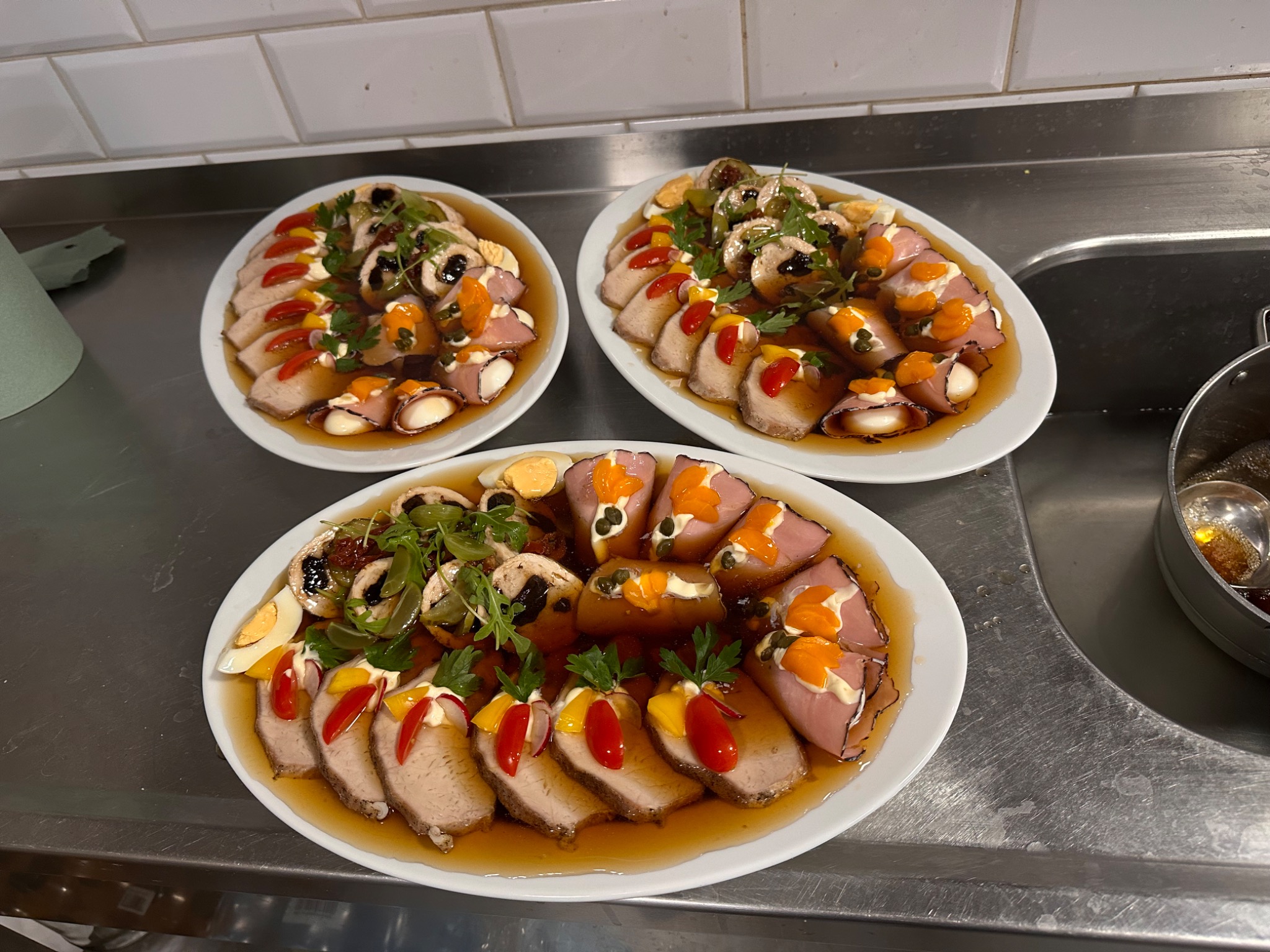 Trzy białe owalne talerze z plastrami pieczonego mięsa w sosie, dekorowane jajkiem, grzybami, zieleniną i kolorowymi warzywami, ustawione na metalowym blacie kuchennym.