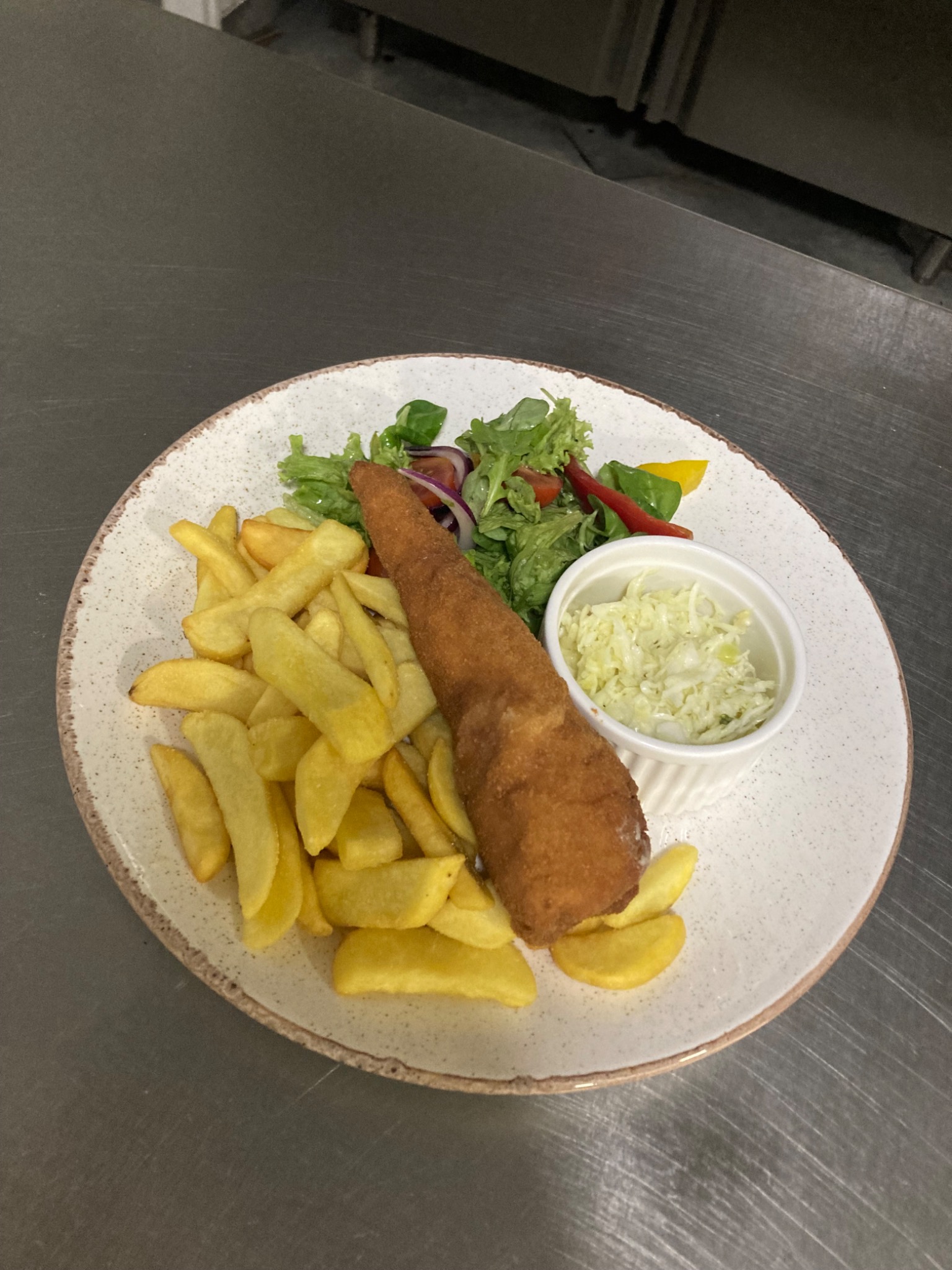 Panierowana ryba podłużna serwowana z frytkami, surówką z kapusty i sałatką warzywną na talerzu z brązową obwódką, ułożona na metalowym blacie.