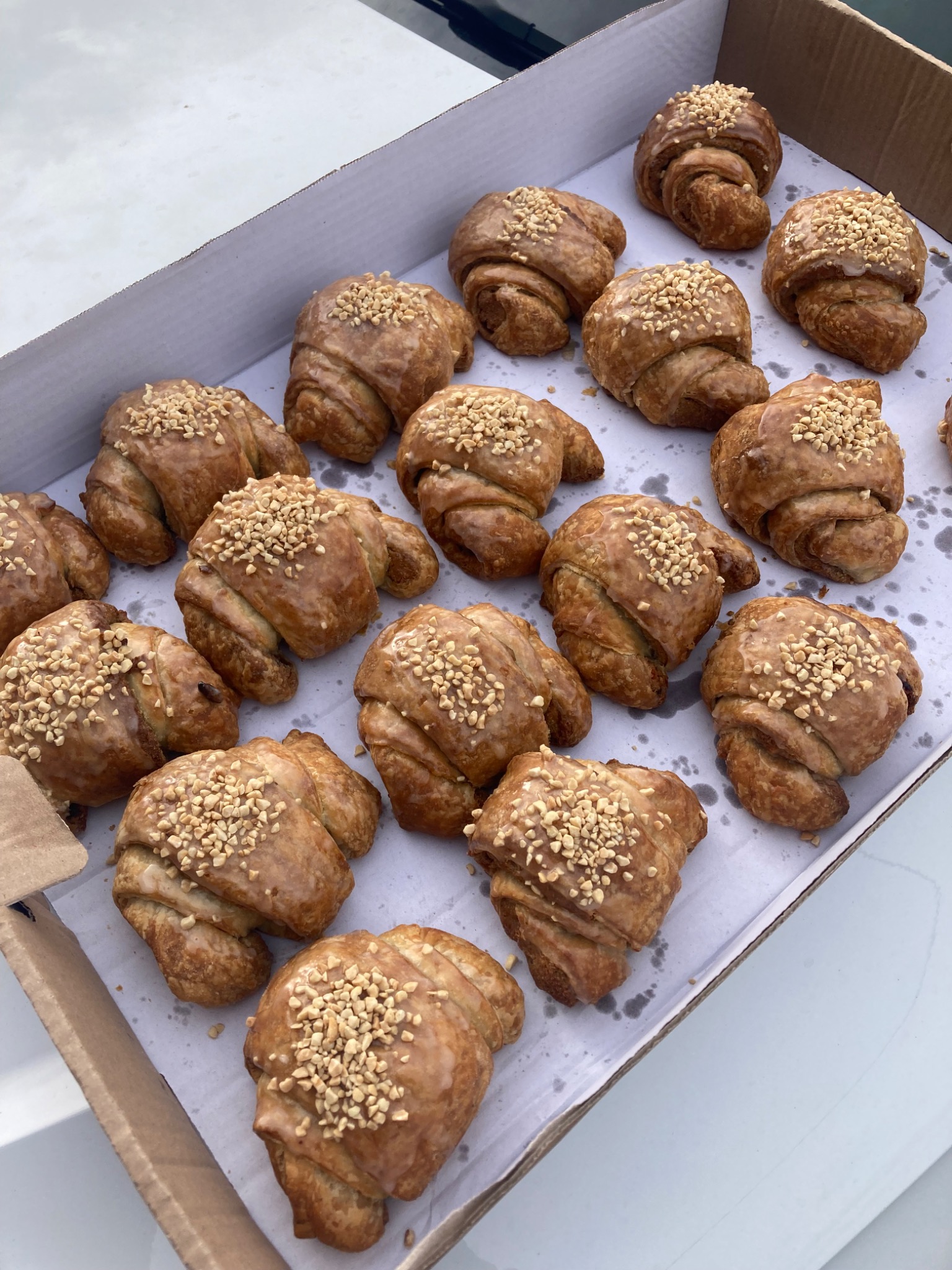 Kartonowe pudełko wypełnione croissantami z błyszczącą polewą i posypką z posiekanych orzechów, ułożonymi na białym papierze.