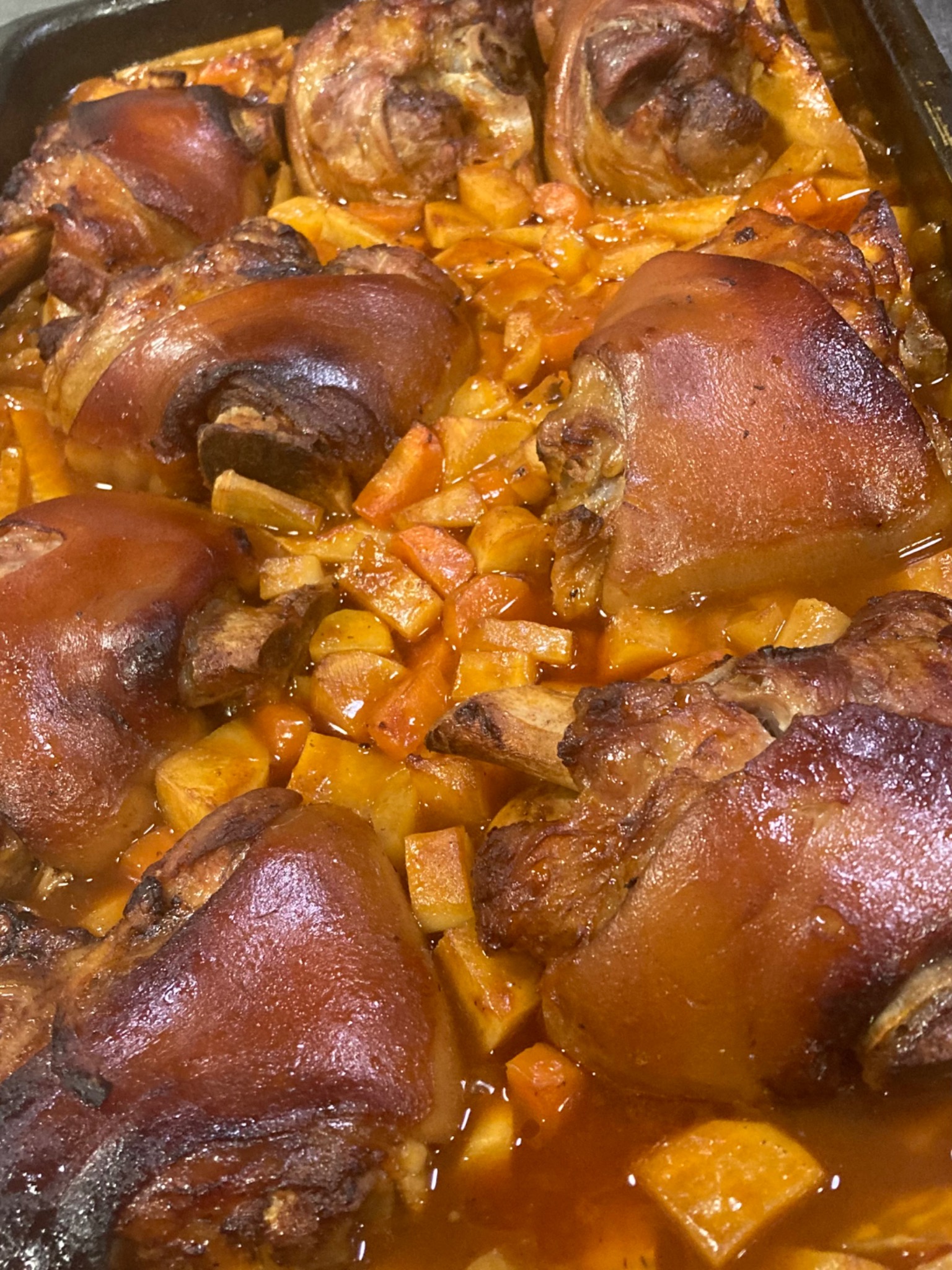 Pieczone golonki wieprzowe z ziemniakami i marchewką w sosie własnym, ułożone w brytfannie, widoczne przypieczone skórki i kawałki warzyw.