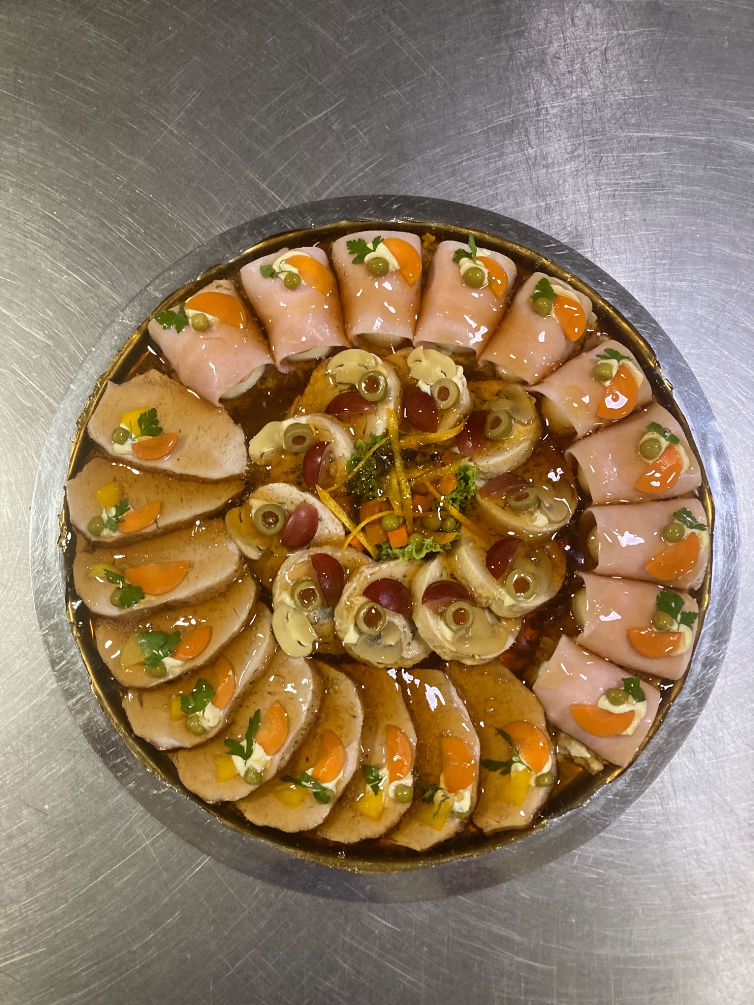 Okrągła taca z różnorodnymi przekąskami: roladki z szynki z dekoracją z moreli i groszku, plastry mięsa w galarecie z warzywami, koreczki z pieczarek i winogron, całość w złotej ramie na metalowym...