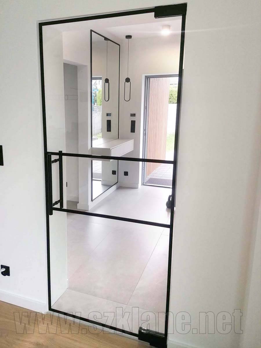 Szklane drzwi loftowe z czarną ramą, widok na przedpokój z lustrem i minimalistycznymi lampami wiszącymi, jasna podłoga z płytek.