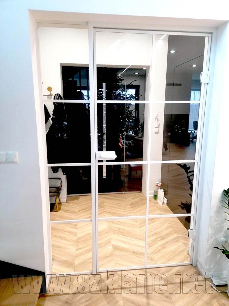 Przeszklone, przesuwne drzwi wewnętrzne z białymi szprosami, odbijające wnętrze z parkietem w jodełkę i choinką, widoczne logo firmy w dolnej części.