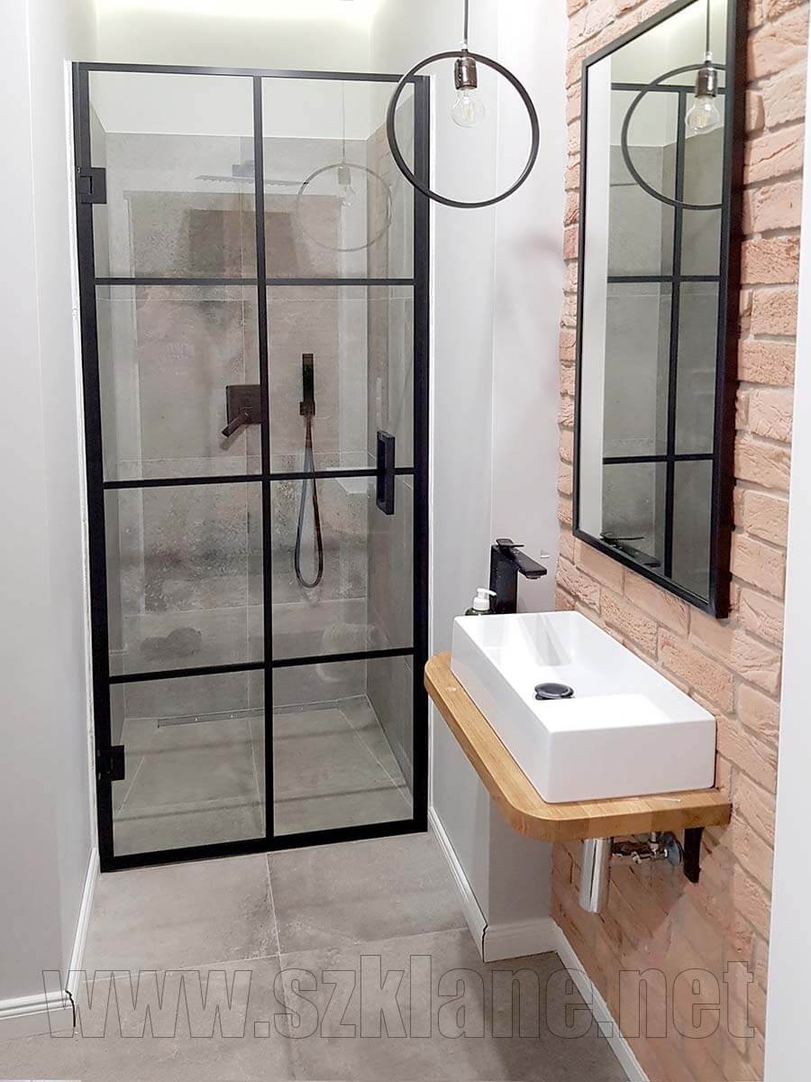 Nowoczesna kabina prysznicowa z czarnymi szprosami, umywalka nablatowa na drewnianym blacie, lustro w czarnej ramie na tle ceglanej ściany, minimalistyczna lampa wisząca.