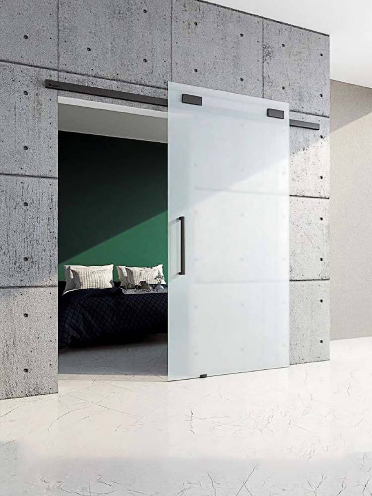 Szklane, przesuwne drzwi w minimalistycznej czarnej ramie, częściowo otwarte, odsłaniające sypialnię z ciemnozieloną ścianą i łóżkiem z czarną narzutą w pomieszczeniu z betonowymi ścianami...