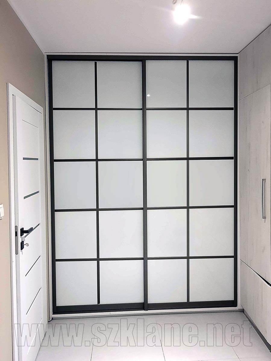 Szafa przesuwna z matowego szkła z czarnymi profilami, podzielona na szesnaście paneli, widoczna z boku, obok białe drzwi i szafka, na dole widoczny adres strony internetowej.