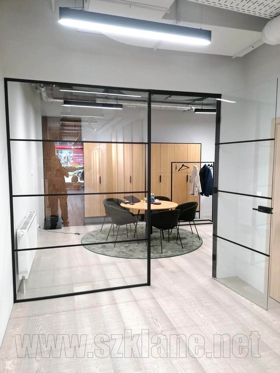 Szklane drzwi loftowe z czarnymi szprosami oddzielające przestrzeń biurową, widoczny okrągły stół z krzesłami i szafy w tle.