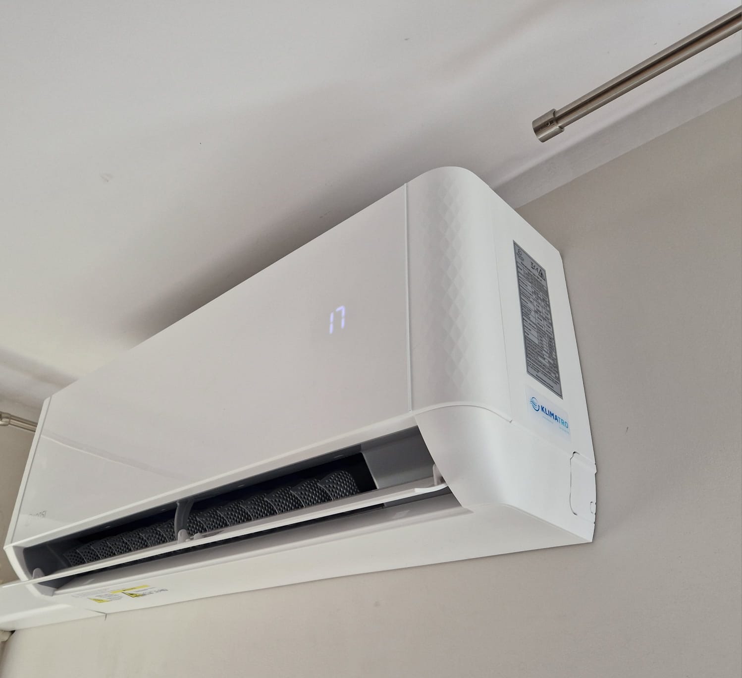 Biały klimatyzator ścienny marki Klimatro zamontowany w pomieszczeniu, wyświetlający temperaturę 17 stopni Celsjusza. Widoczny fragment sufitu i karnisza.