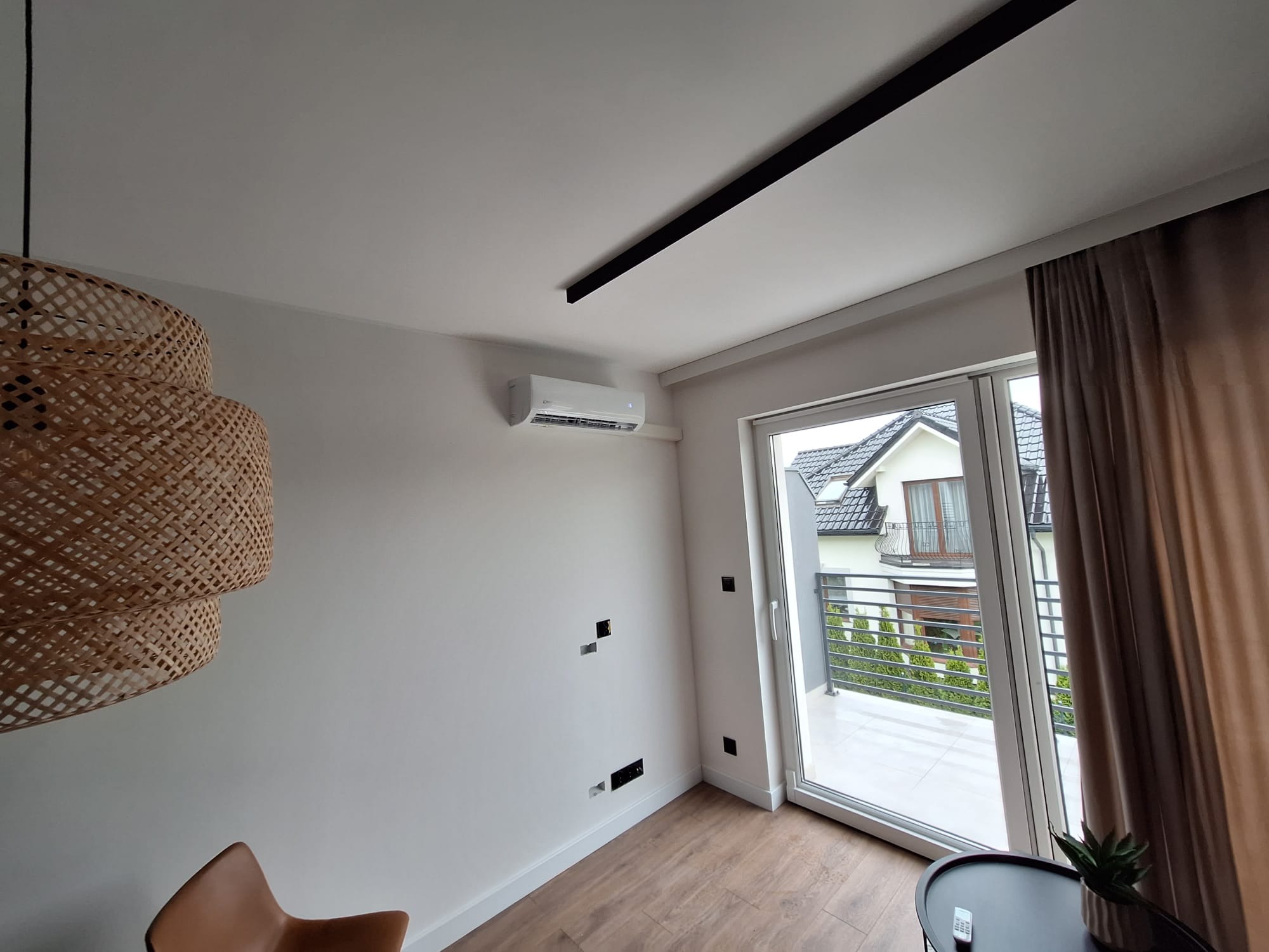 Biały klimatyzator na ścianie w jasnym pokoju z drewnianą podłogą, widok na balkon i dom przez okno balkonowe, nowoczesna lampa z wikliny.