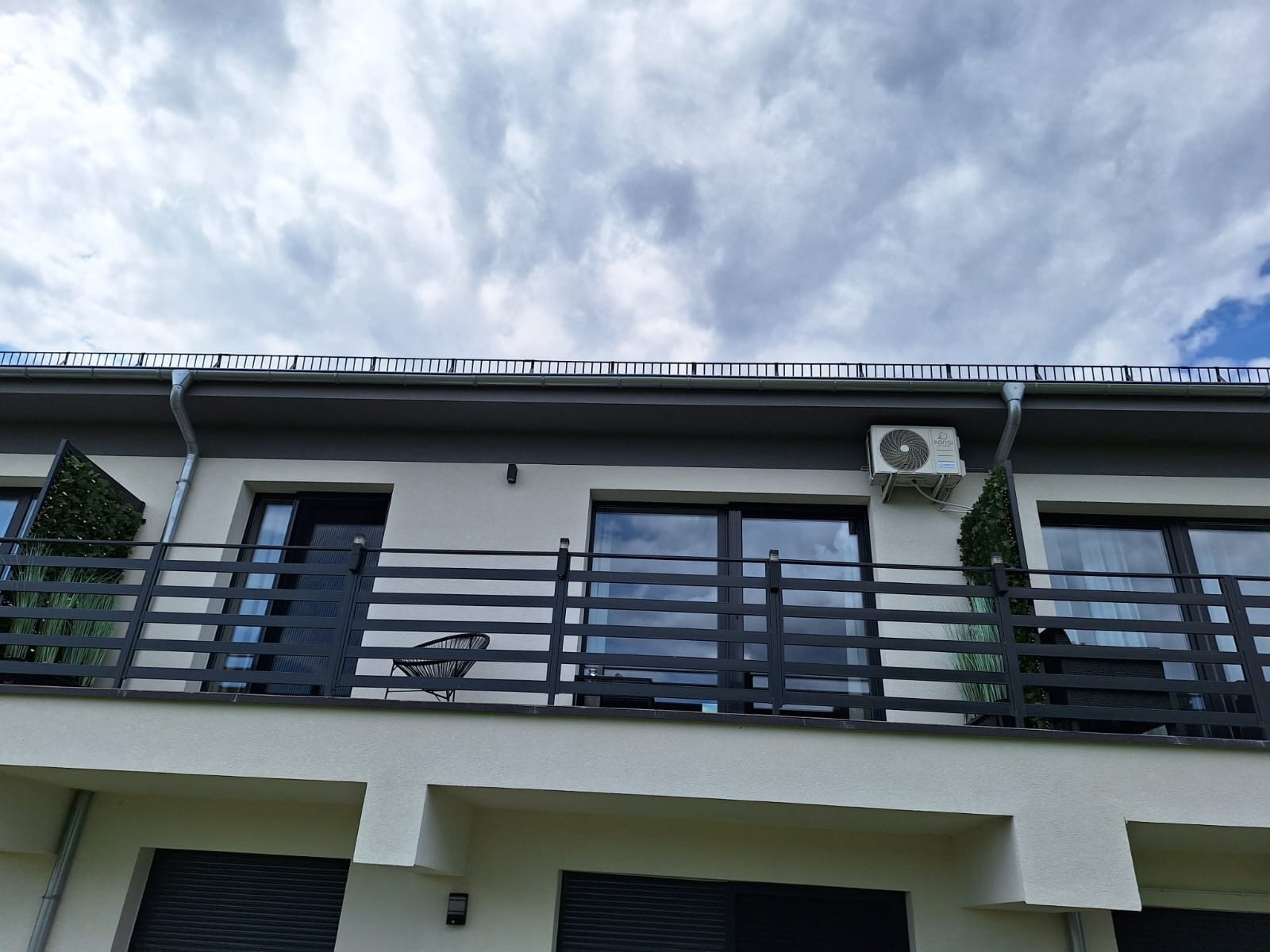 Biały budynek z balkonem, na którym stoi czarne krzesło i stolik. Na ścianie zamontowana jednostka zewnętrzna klimatyzacji marki Rotenso. Widok z dołu na pochmurne niebo.