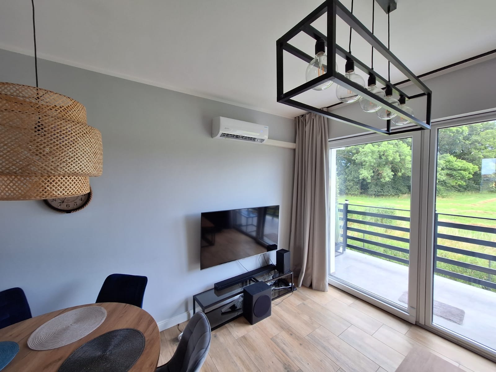 Wnętrze salonu z zamontowaną klimatyzacją ścienną, widok na balkon przez duże okno przesuwne, nowoczesna lampa sufitowa z żarówkami Edisona, okrągły stół z podkładkami i krzesłami.