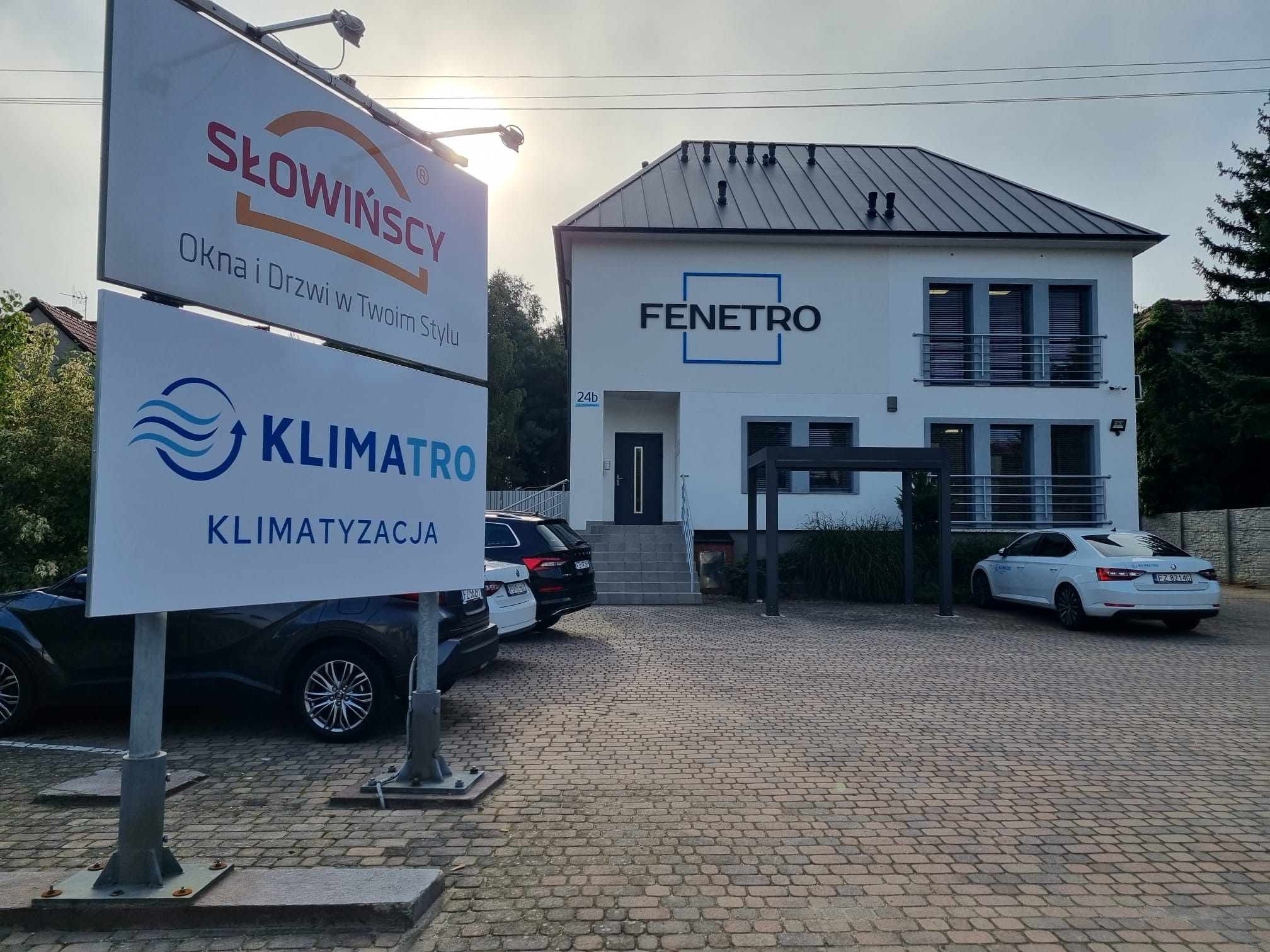 Budynek firmy z szyldem reklamowym oferującym okna, drzwi i systemy chłodzenia, zaparkowane samochody na brukowanym podjeździe.