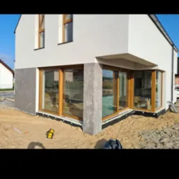 Nowoczesny dom jednorodzinny z dużymi oknami narożnymi, szarą dolną częścią elewacji i jasną g&oacute;rną, w trakcie prac wykończeniowych wok&oacute;ł budynku, widoczny niwelator laserowy.