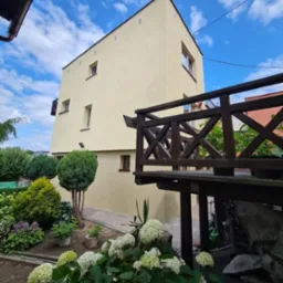 Dwupiętrowy dom z beżową elewacją i ciemnym dachem, widziany z poziomu ogrodu z kwitnącymi hortensjami i drewnianą balustradą tarasu.