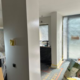 MACH-BUD Daniel Machajewski - Wnętrze w trakcie malowania: ściany w odcieniach szarości i bieli, zabezpieczona folią okno i podłoga z taśmą malarską, widoczne puszki z farbą i narzędzia.