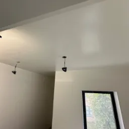 Wnętrze po malowaniu na biało, widoczne gładkie ściany, sufit z wystającymi przewodami od oświetlenia i okno z widokiem na drzewa.