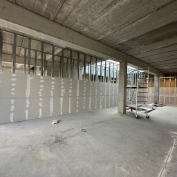 MACH-BUD Daniel Machajewski - Wnętrze biurowe w trakcie prac wykończeniowych: widoczne metalowe profile pod ścianki działowe, płyty gipsowo-kartonowe, surowy betonowy strop i drabina na kółkach.