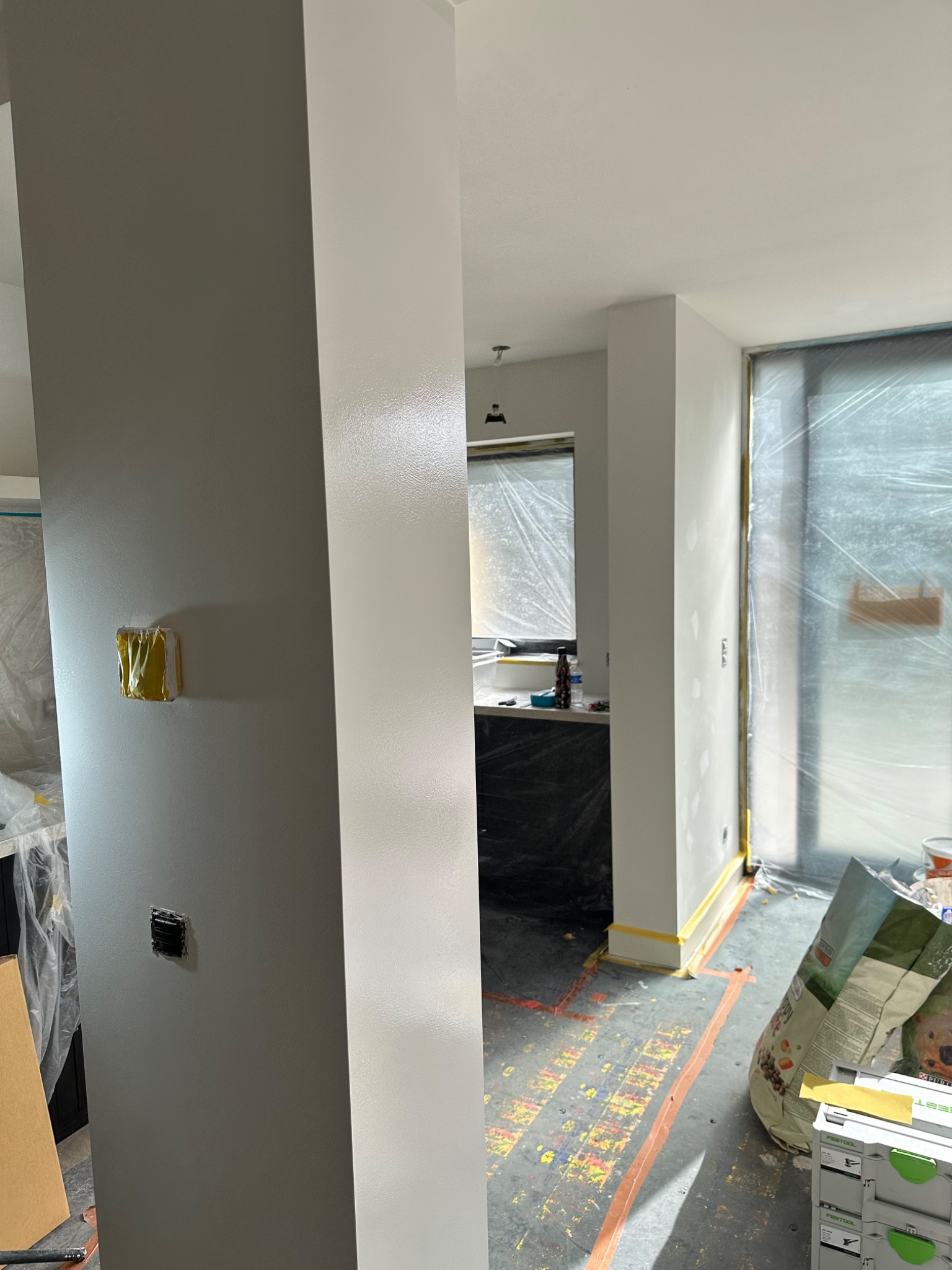 Wnętrze w trakcie malowania: ściany w odcieniach szarości i bieli, zabezpieczona folią okno i podłoga z taśmą malarską, widoczne puszki z farbą i narzędzia.