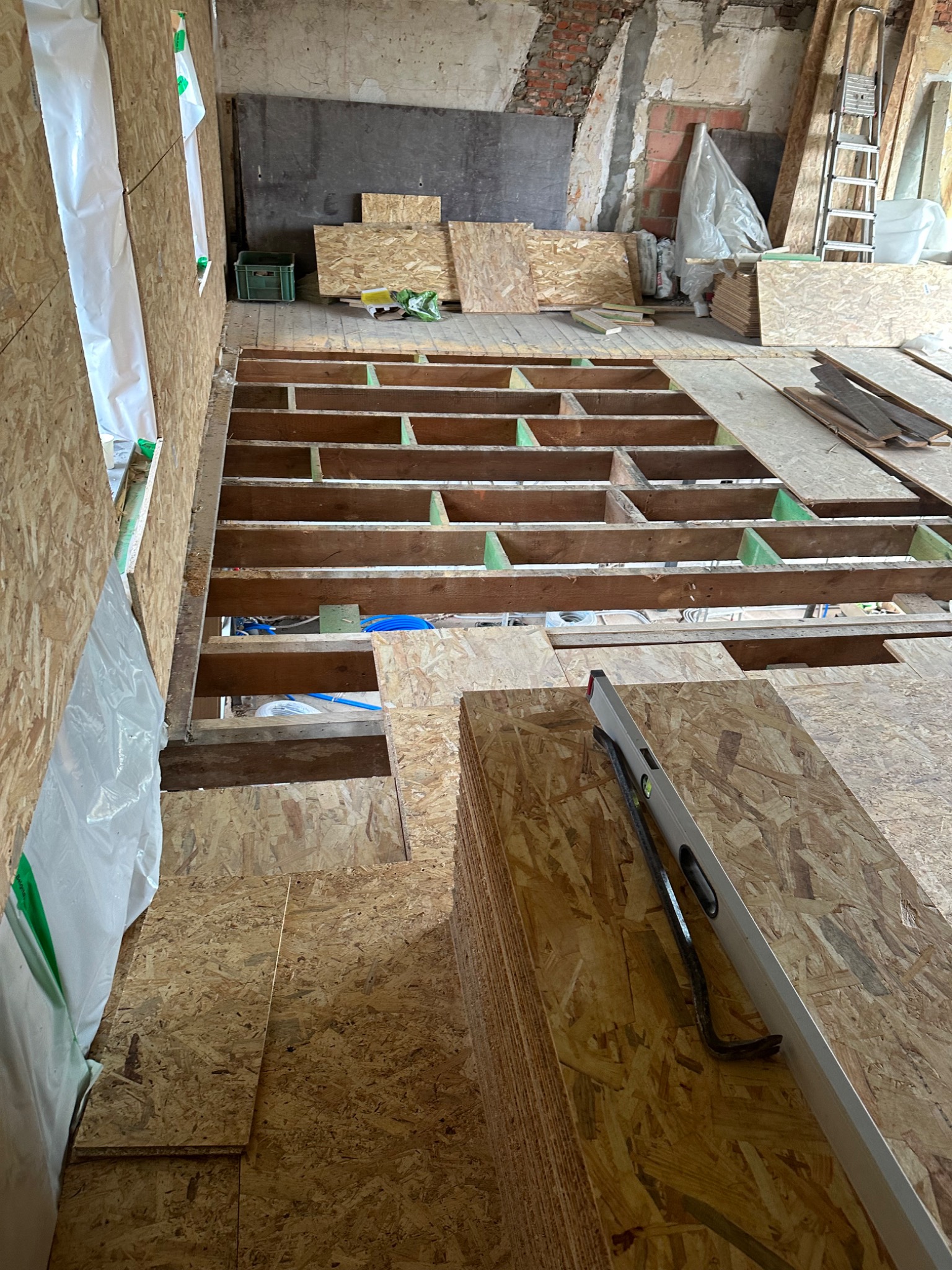 Konstrukcja drewniana podłogi na legarach z widocznymi belkami i płytami OSB, poziomica i łom na stosie płyt, surowe ściany w tle.