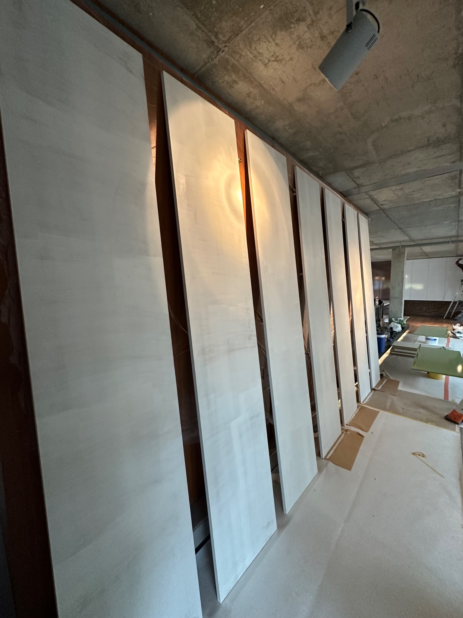 Świeżo pomalowane na biało panele drzwiowe lub ścienne, ustawione pionowo do wyschnięcia w surowym wnętrzu z betonowym sufitem i widocznym oświetleniem punktowym, zabezpieczona podłoga.