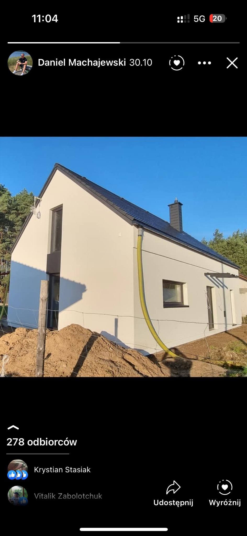 Jasna elewacja nowo wybudowanego domu jednorodzinnego z ciemną dachówką, widoczna z boku, z żółtą rurą spustową i stertą ziemi w pobliżu, na tle drzew i błękitnego nieba.