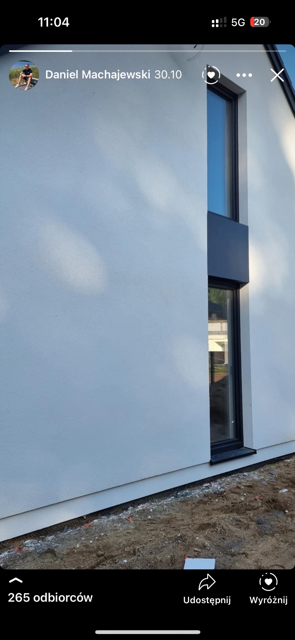 Biała elewacja budynku z oknem o czarnej ramie, widoczne odbicie nieba w szybie, cień drzewa na ścianie.