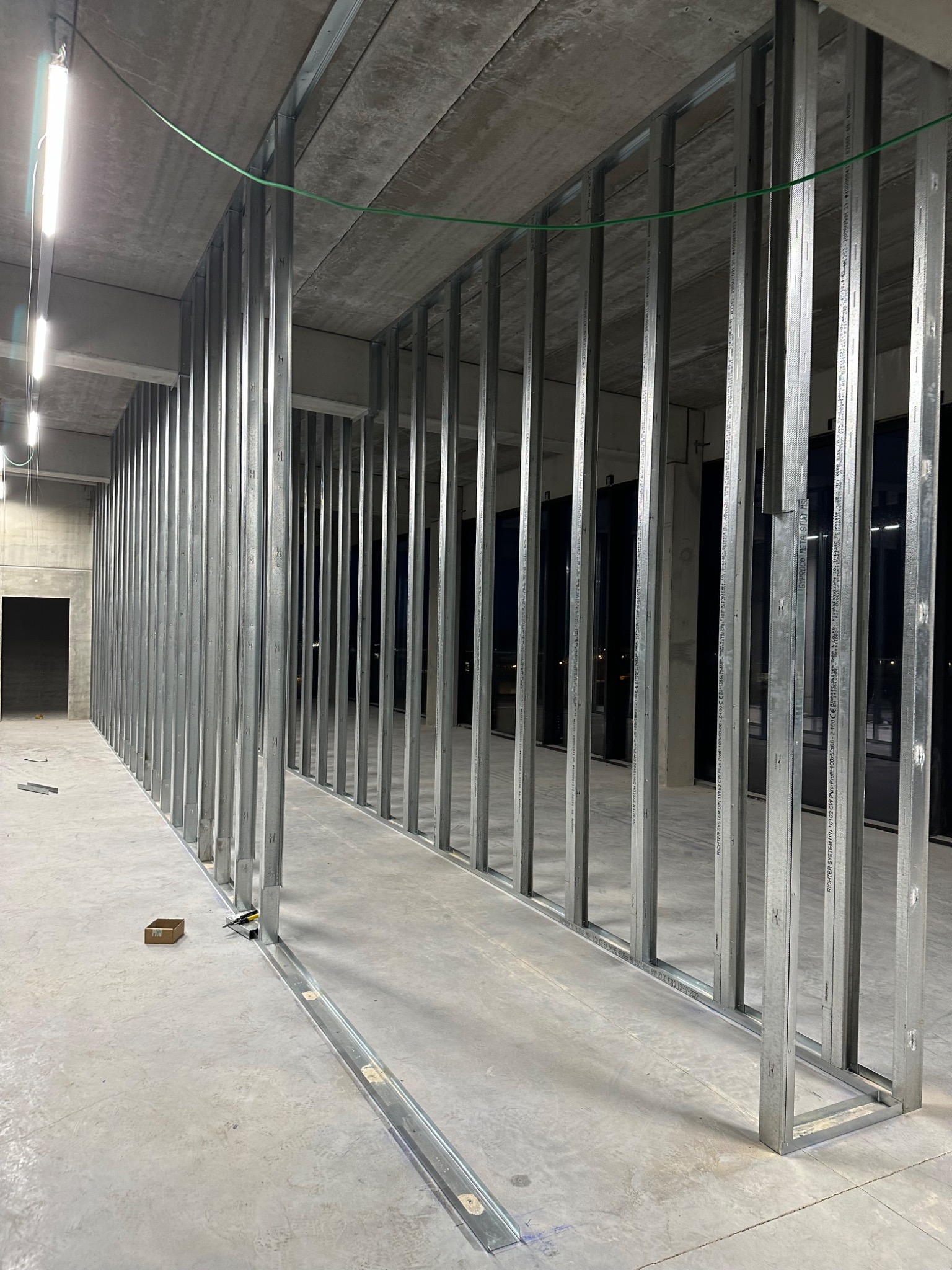 Konstrukcja szkieletowa z metalowych profili w surowym stanie, widoczne profile pionowe i poziome, betonowa podłoga i sufit, przygotowanie do montażu płyt gipsowo-kartonowych, mały karton...