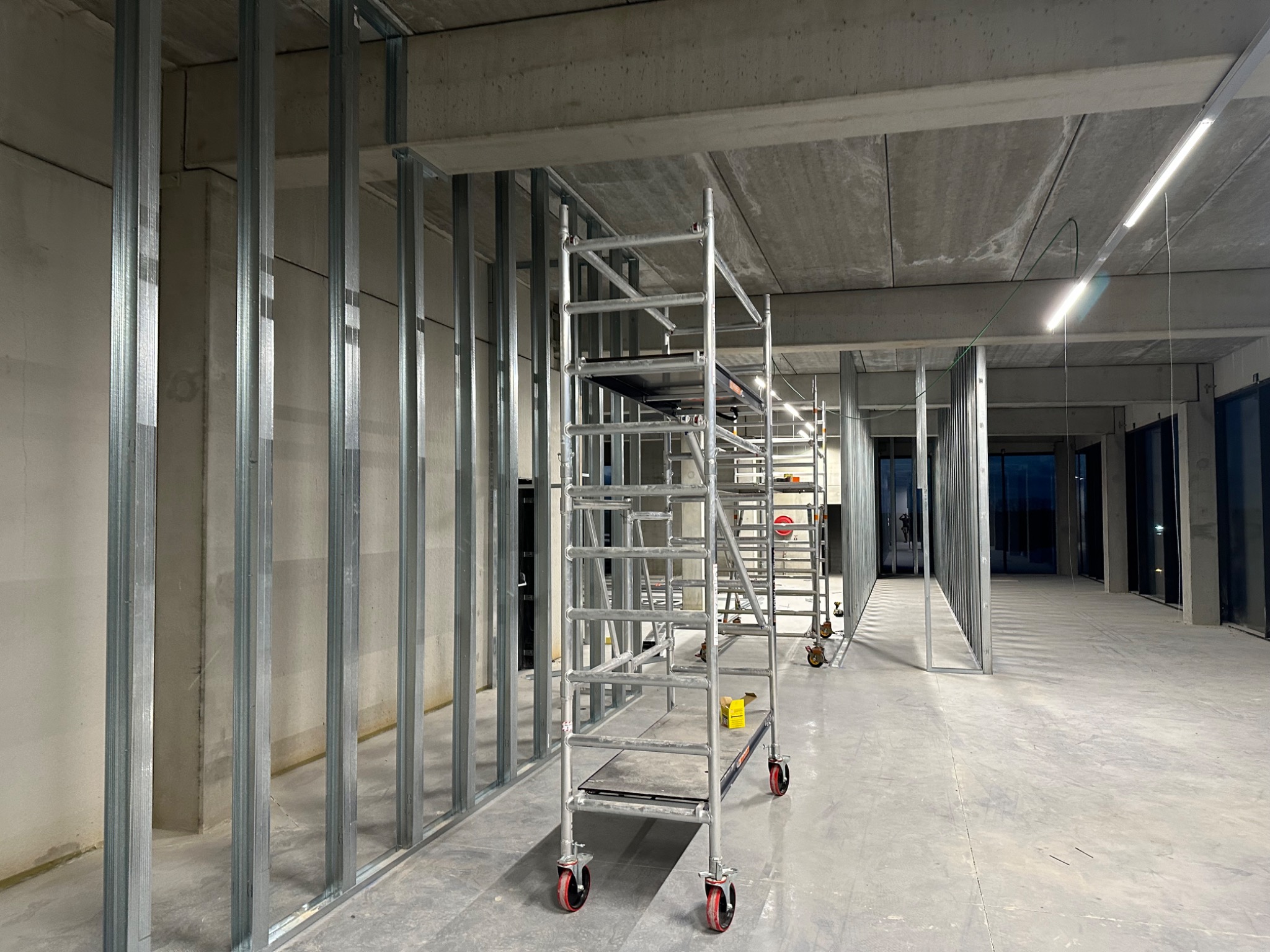 Wnętrze komercyjne w trakcie wykańczania: metalowe profile ścian działowych, rozstawione rusztowania na kółkach, surowy betonowy strop z oświetleniem liniowym.