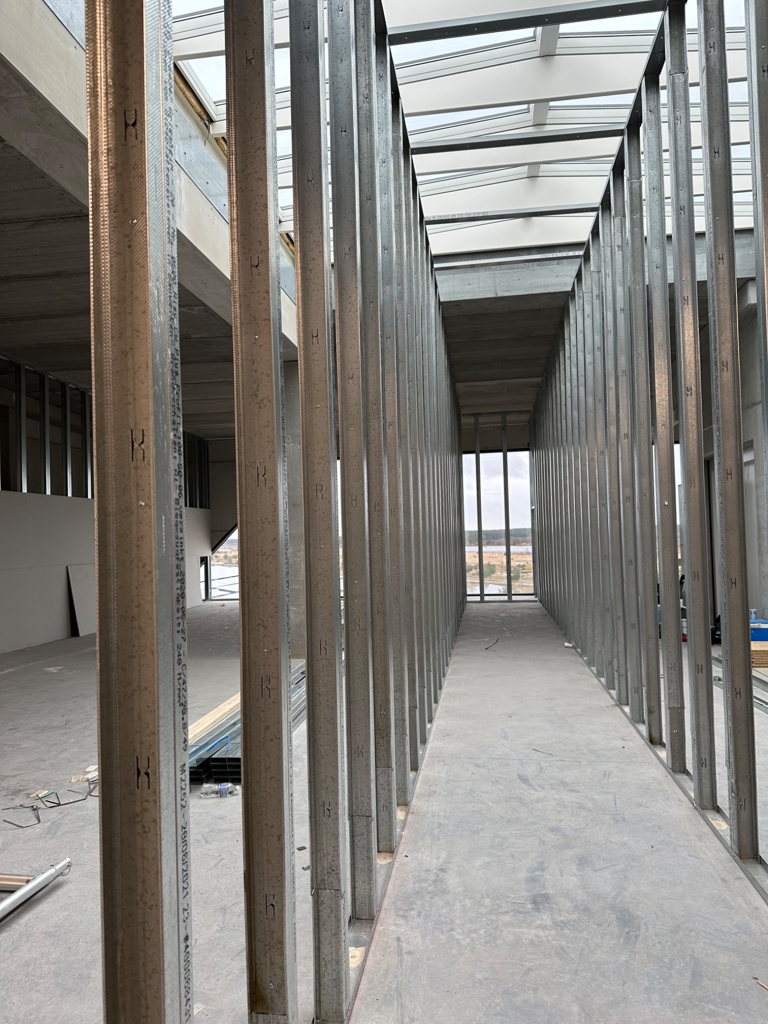 Wnętrze w trakcie budowy z widocznymi metalowymi profilami ścian działowych, surowy betonowy sufit z oknami dachowymi i widokiem na krajobraz w oddali.