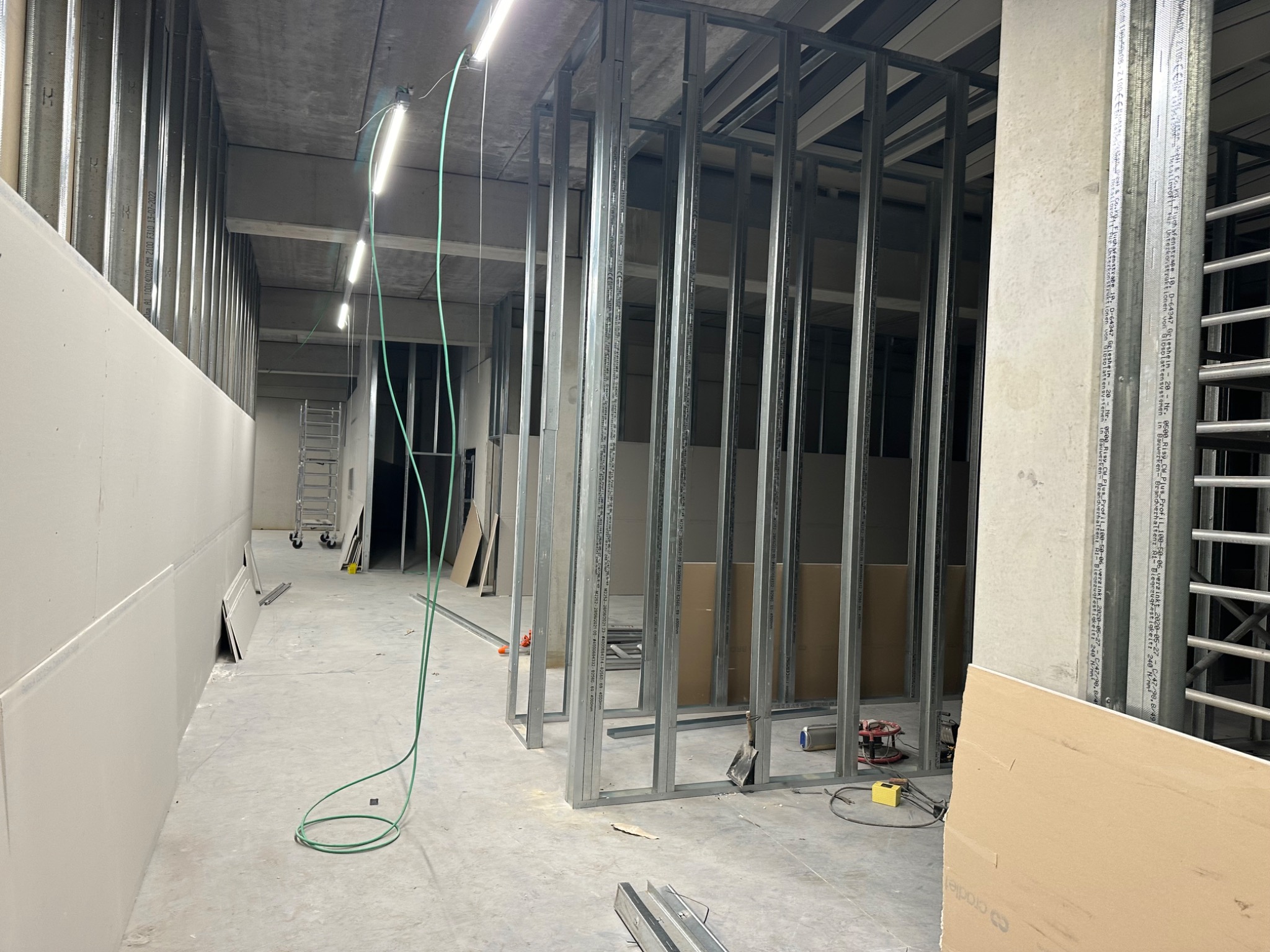 Wnętrze w trakcie prac budowlanych: metalowa konstrukcja ścian działowych, częściowo obłożona płytami gipsowo-kartonowymi, widoczne narzędzia i materiały budowlane, surowy betonowy strop...