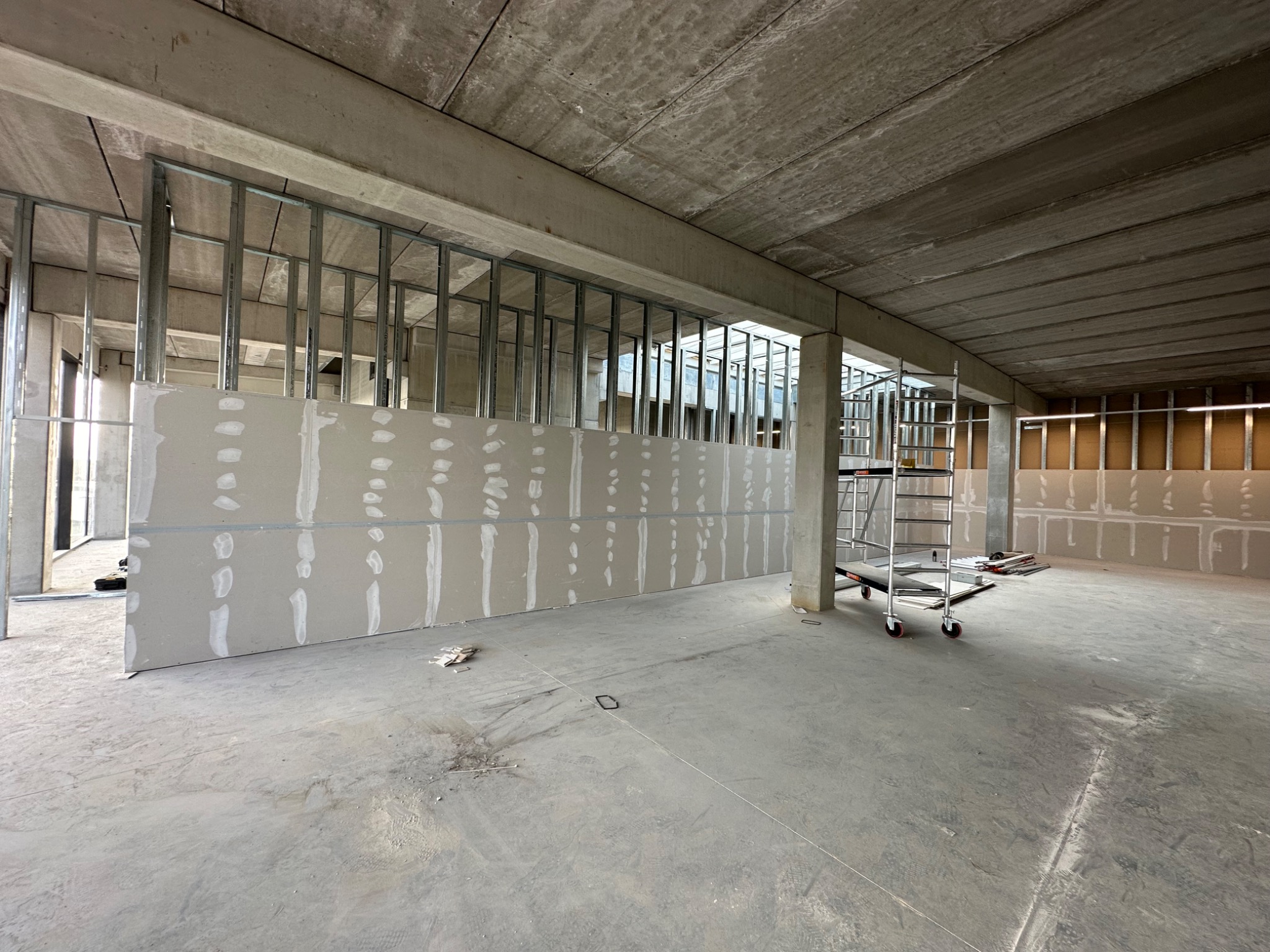 Wnętrze biurowe w trakcie prac wykończeniowych: widoczne metalowe profile pod ścianki działowe, płyty gipsowo-kartonowe, surowy betonowy strop i drabina na kółkach.