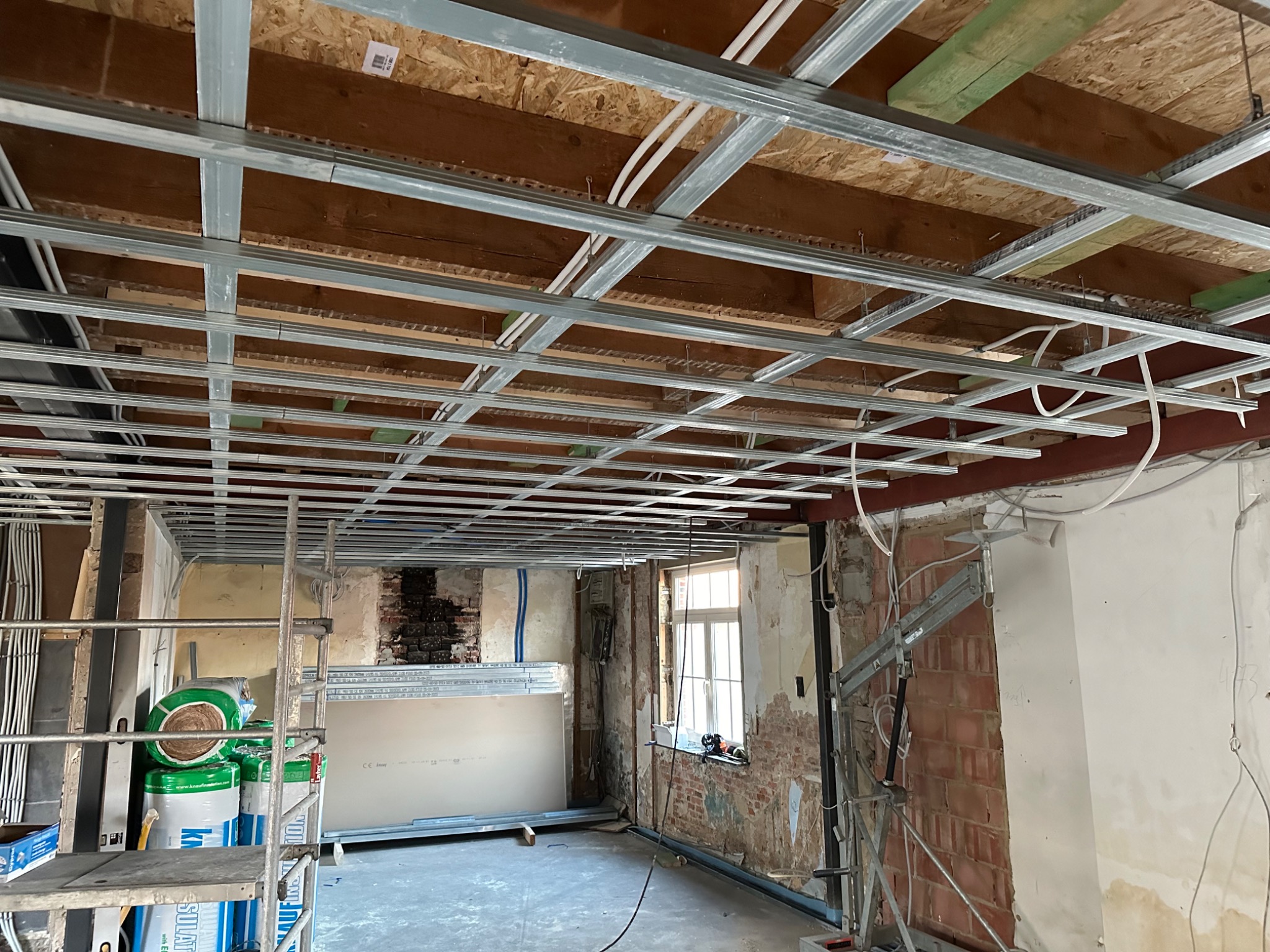 Wnętrze w trakcie montażu metalowej konstrukcji sufitu podwieszanego, widoczne przewody elektryczne, płyty gipsowo-kartonowe oparte o ścianę oraz materiały izolacyjne na rusztowaniu.