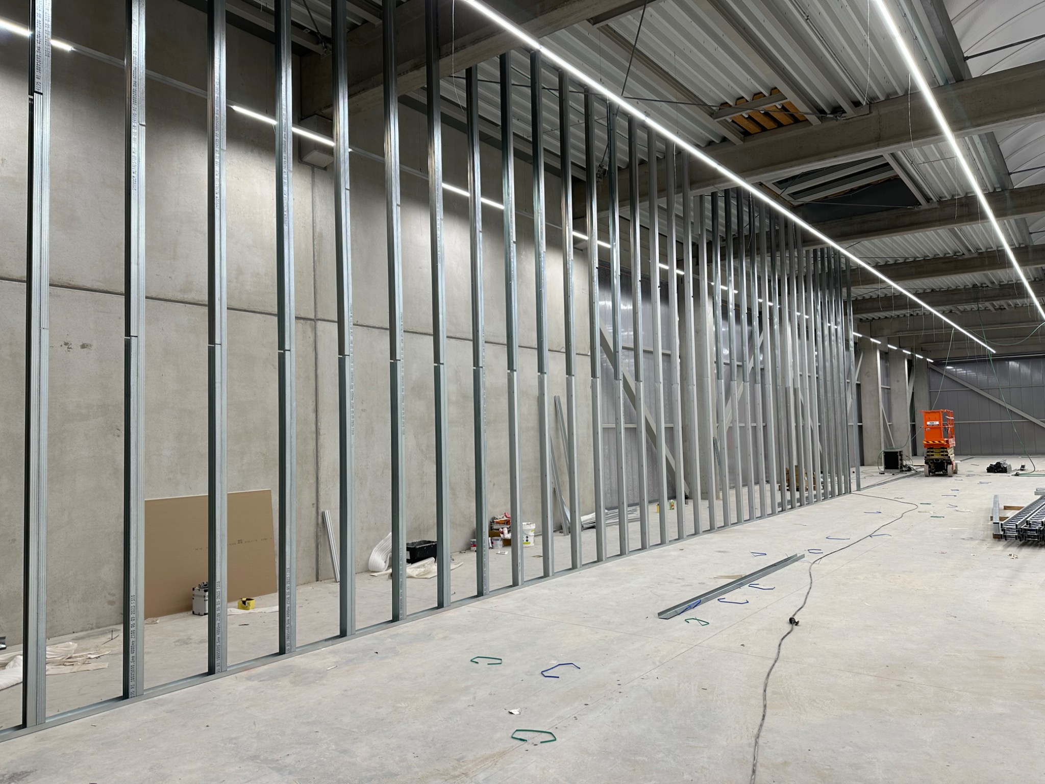 Montaż metalowej konstrukcji szkieletowej ścian działowych w hali przemysłowej, widoczna płyta gipsowo-kartonowa, narzędzia i podnośnik koszowy w tle.