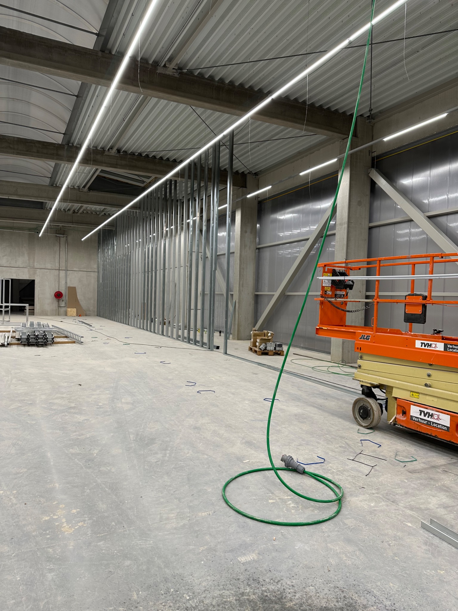 Wnętrze hali produkcyjnej z metalową konstrukcją ścian działowych w trakcie montażu, oświetleniem LED i podnośnikiem koszowym JLG.