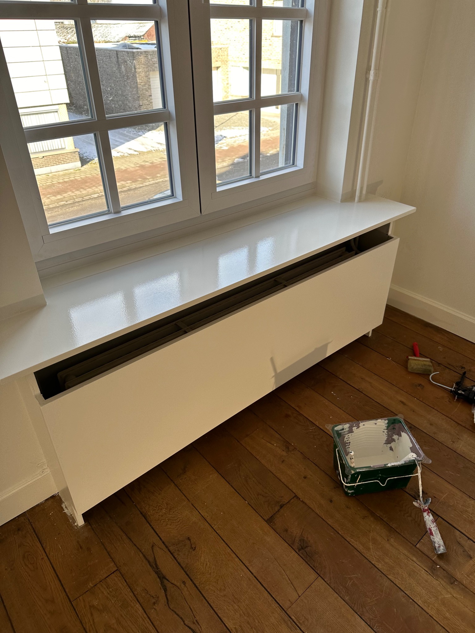 Biała, świeżo pomalowana obudowa grzejnika pod oknem, widoczne ślady farby na podłodze z drewnianych desek, narzędzia malarskie obok, w tle budynki za oknem.