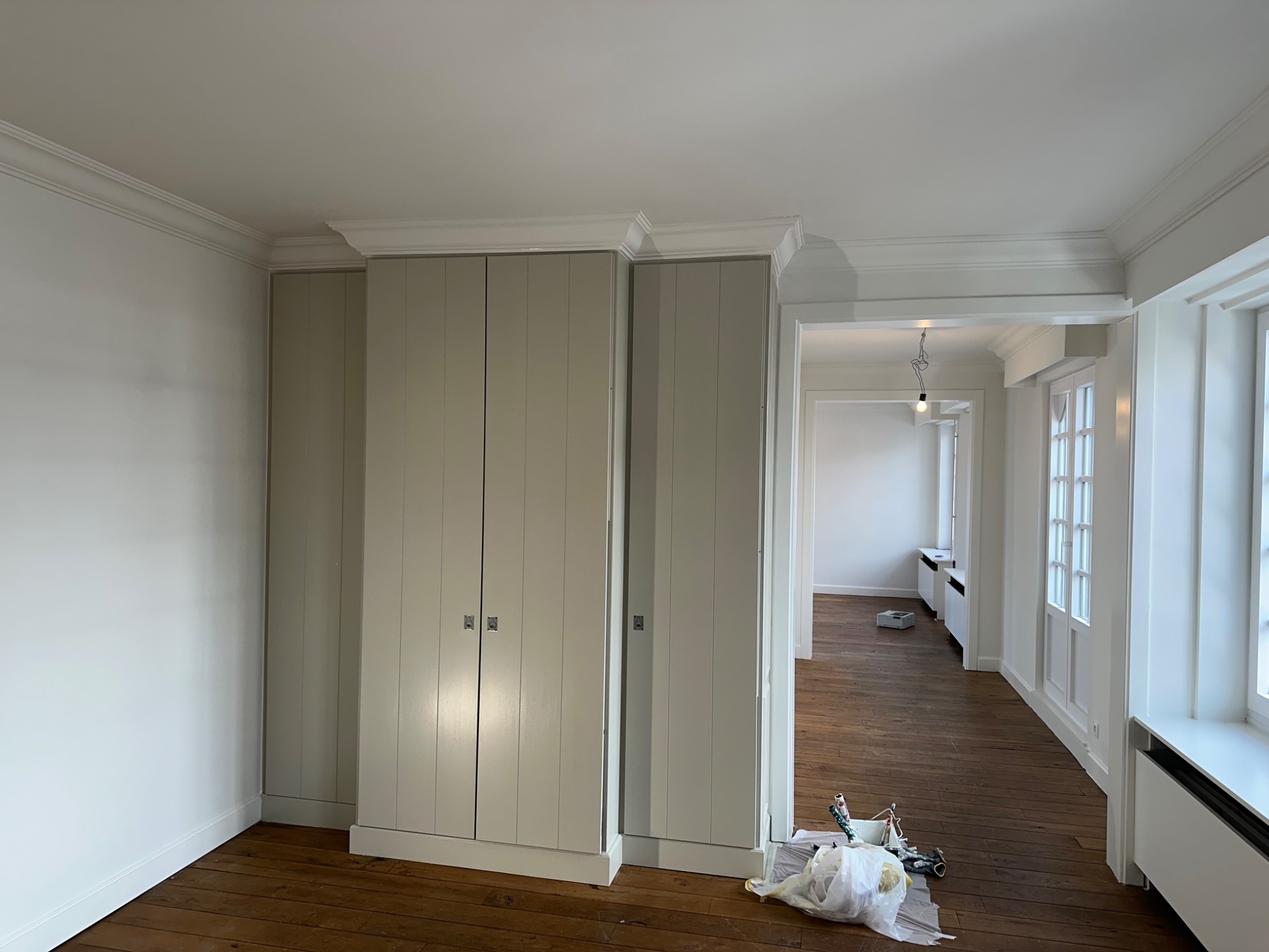 Wnętrze po malowaniu: odświeżone ściany w odcieniach bieli, zabudowane szafy w kolorze beżowym z subtelnymi srebrnymi uchwytami, drewniana podłoga i widoczne akcesoria malarskie.